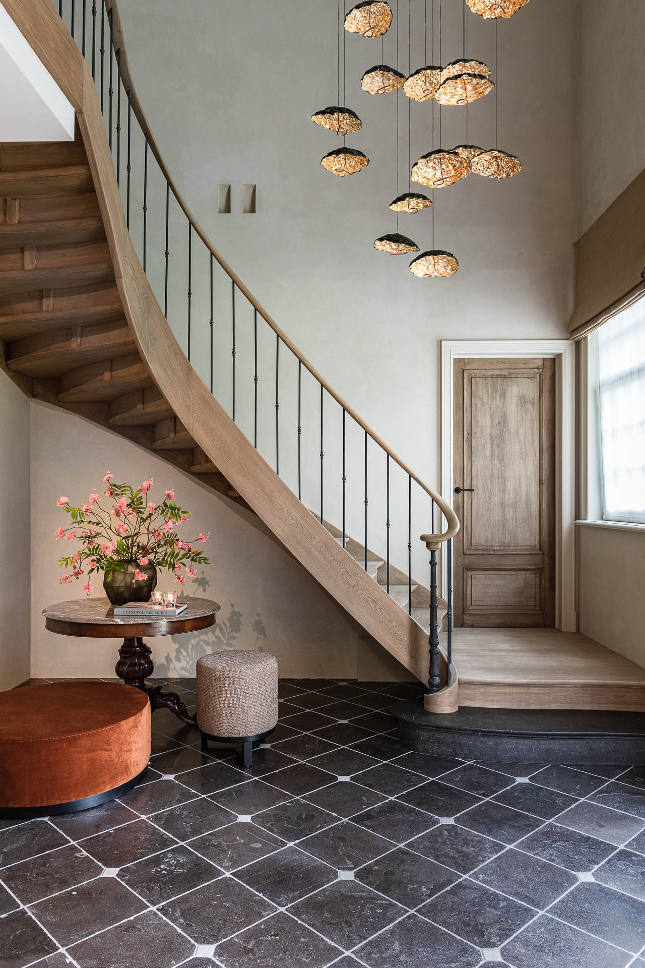 Een stijlvol ingerichte hal met een gebogen houten trap en slanke metalen balustrade; de ruimte heeft een donkere tegelvloer met patroon, een houten deur, een groot raam, een ronde tafel met bloemen en een cluster van moderne hanglampen aan het hoge plafond.