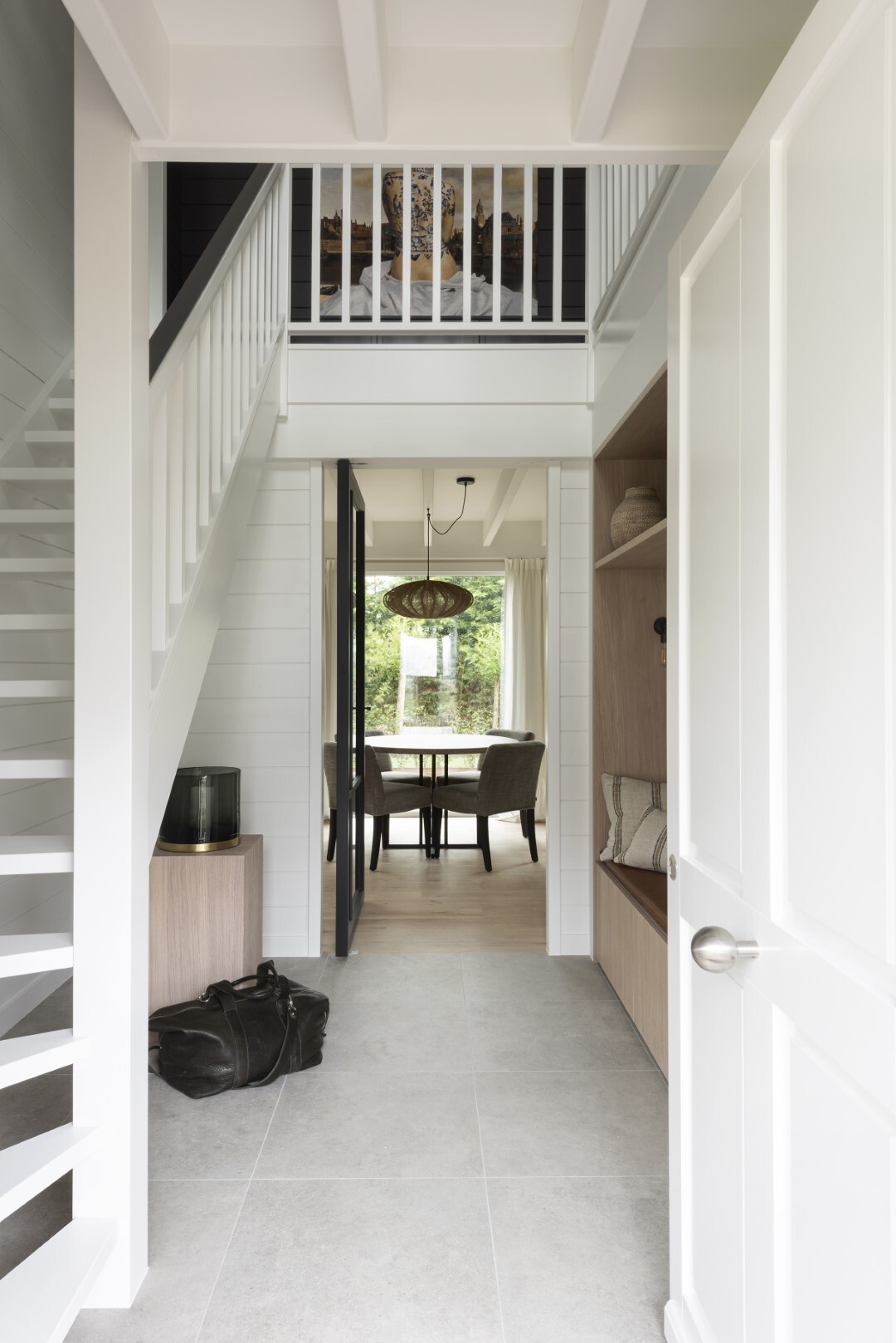 Witte inkomhal met trap links, opbergmeubel rechts en doorkijk naar een eetruimte met grote ramen en uitzicht op de tuin.