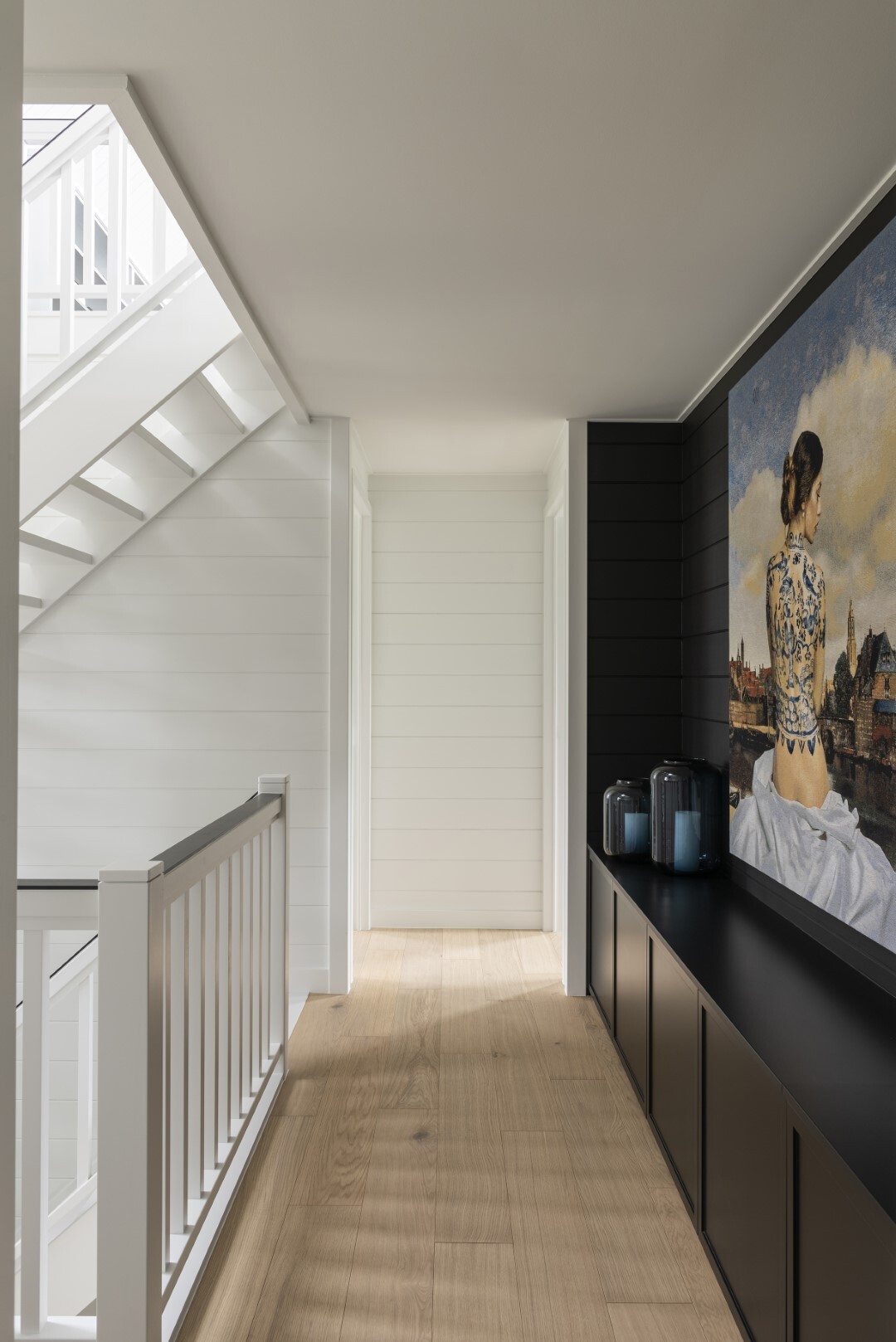Lichte gang met houten vloer, zwarte kastenwand en groot schilderij van een vrouw, naast een witte trap met balustrade.