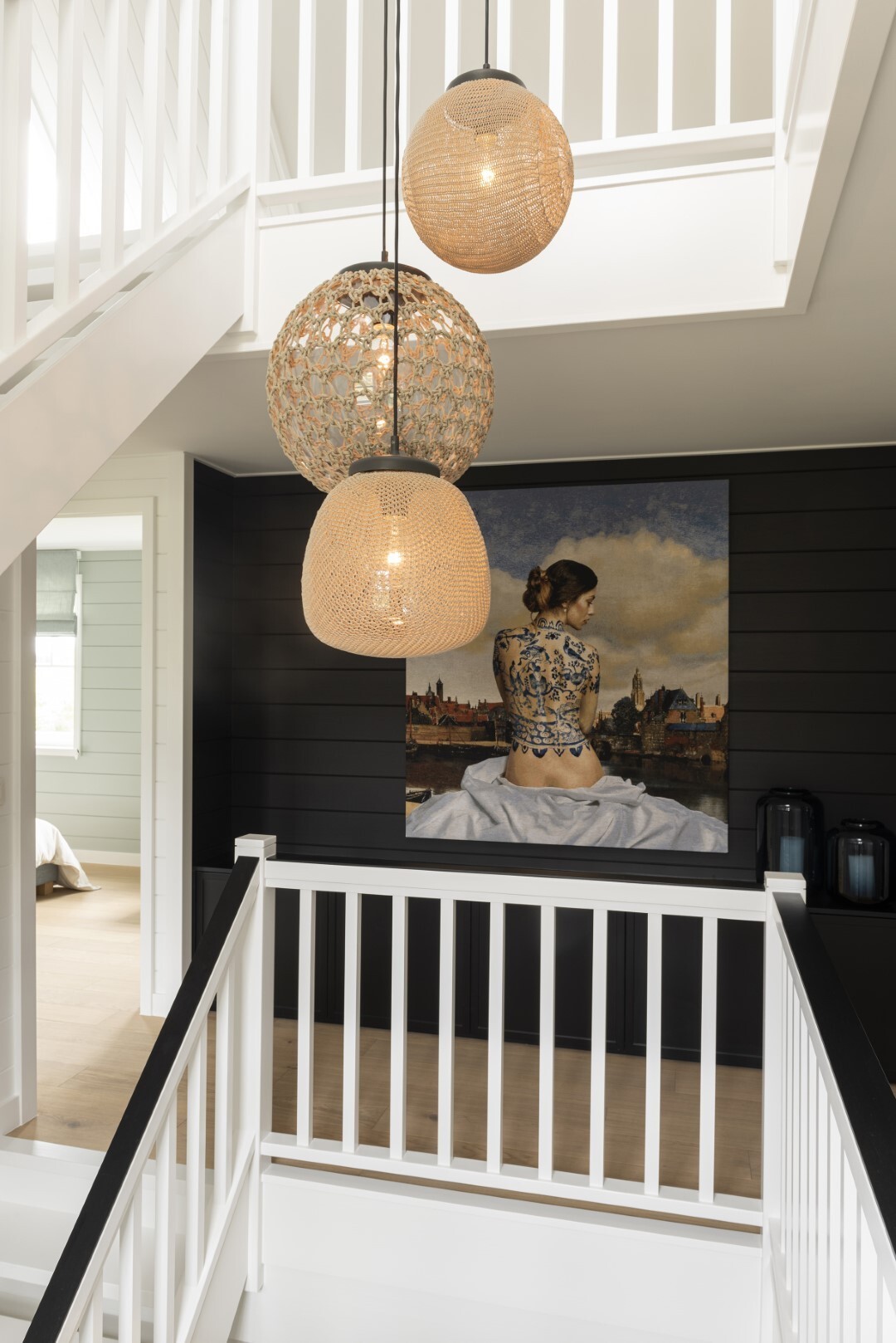 Witte trap en balustrade rond een vide met drie rotan hanglampen, en een schilderij van een vrouw aan een zwarte houten muur.