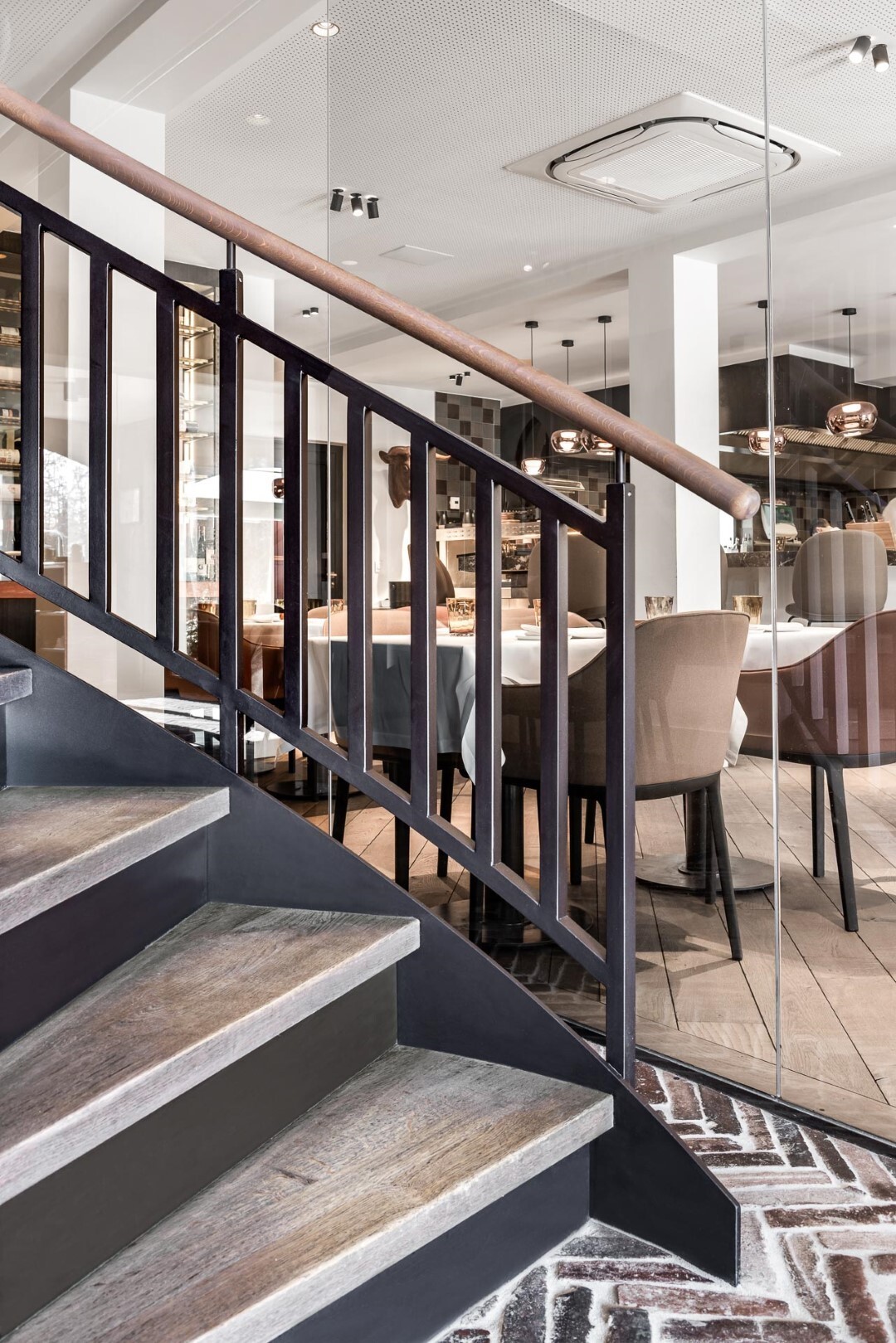 Trap met zwarte balustrade en houten leuning, naast een glazen wand met zicht op een elegante eetruimte met beige stoelen en gedekte tafels.
