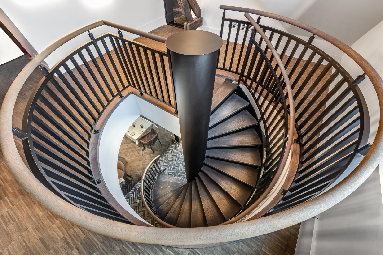 Een elegante spiraaltrap met zwarte treden, houten leuning en een centrale zwarte kolom, gezien van bovenaf met zicht op de verdieping eronder.