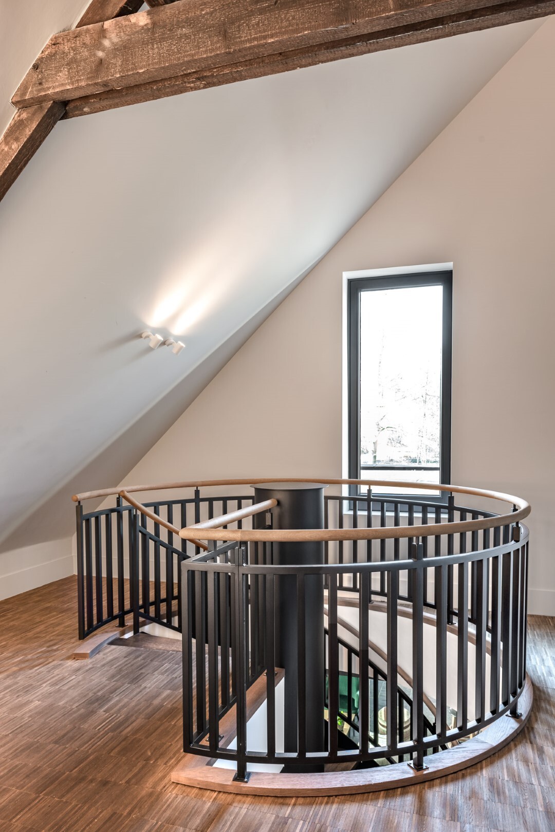 Bovenverdieping met ronde trapopening en zwarte balustrade, onder een schuin plafond met houten draagbalk en een smal verticaal raam.