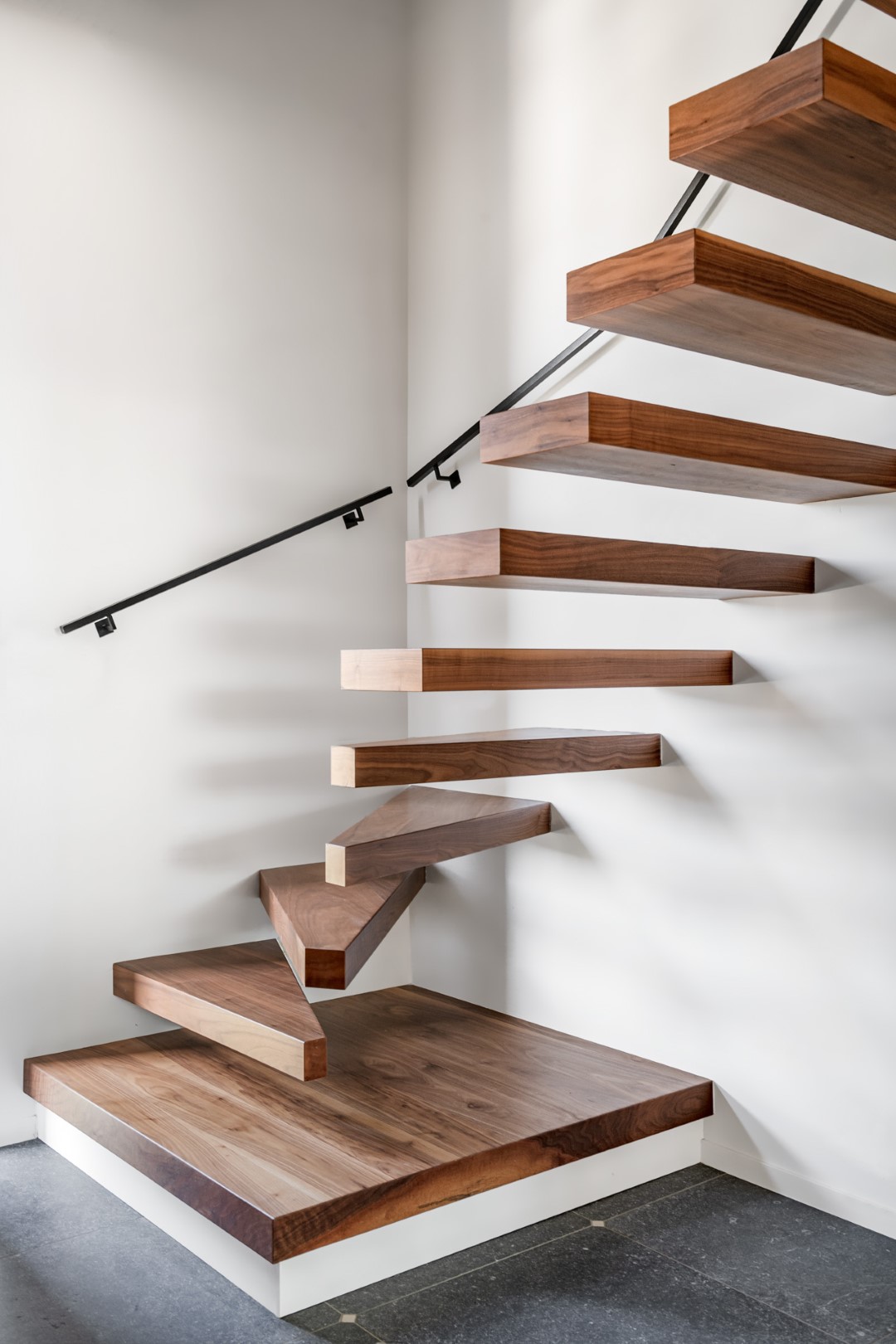 Close-up van een moderne zwevende houten trap met donkerbruine treden, witte muur, zwarte leuning en een platform van hetzelfde hout boven een grijze tegelvloer.
