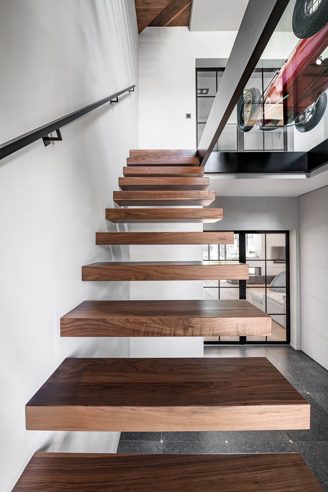 Een moderne zwevende trap met dikke houten treden en een zwarte balustrade, met zicht op een rode speelgoedauto bovenaan.