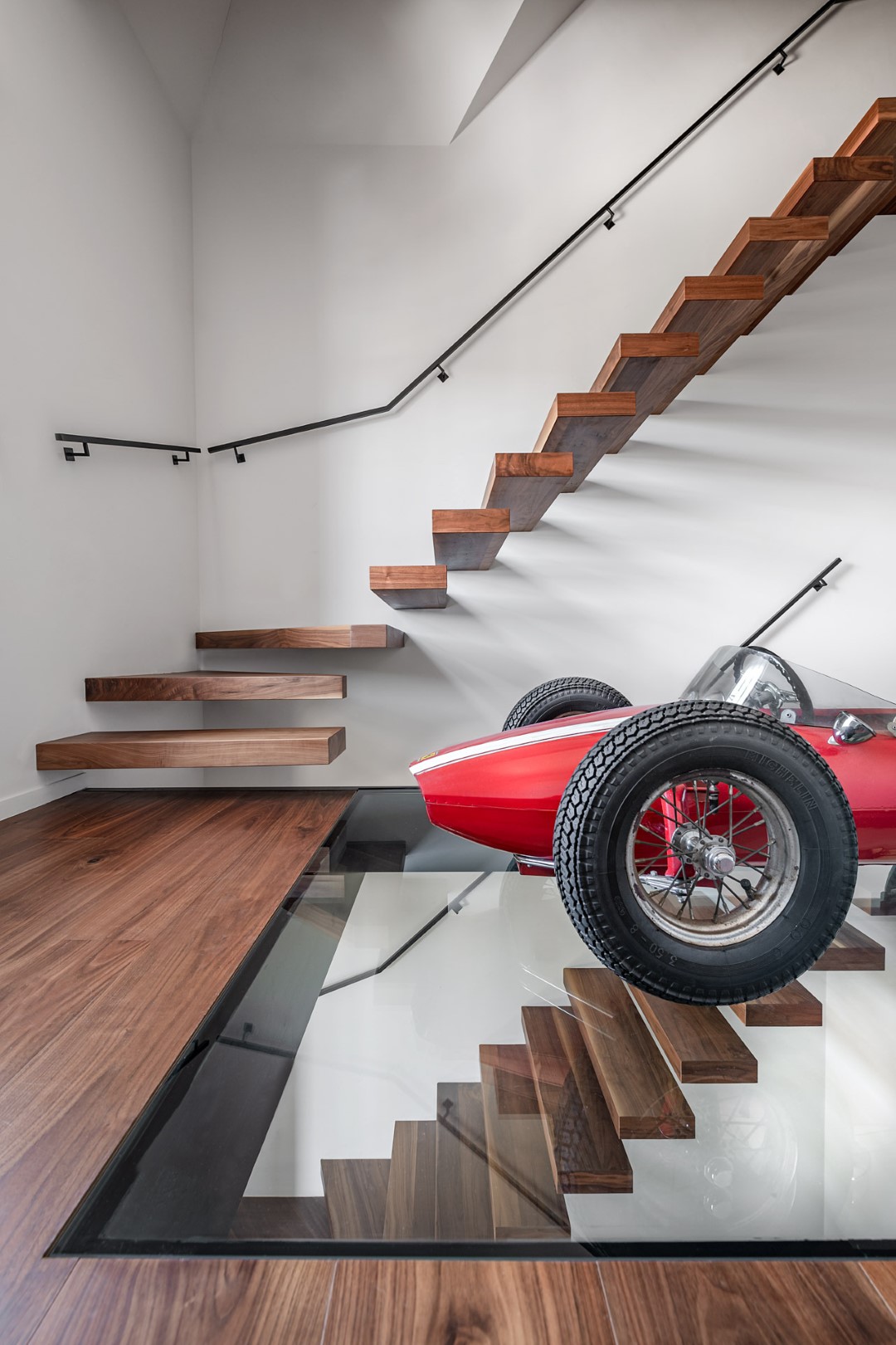Zwevende houten trap langs een witte muur, met onderaan een glazen vloer waaronder de trap zichtbaar is en een rode speelgoedracewagen erbovenop.