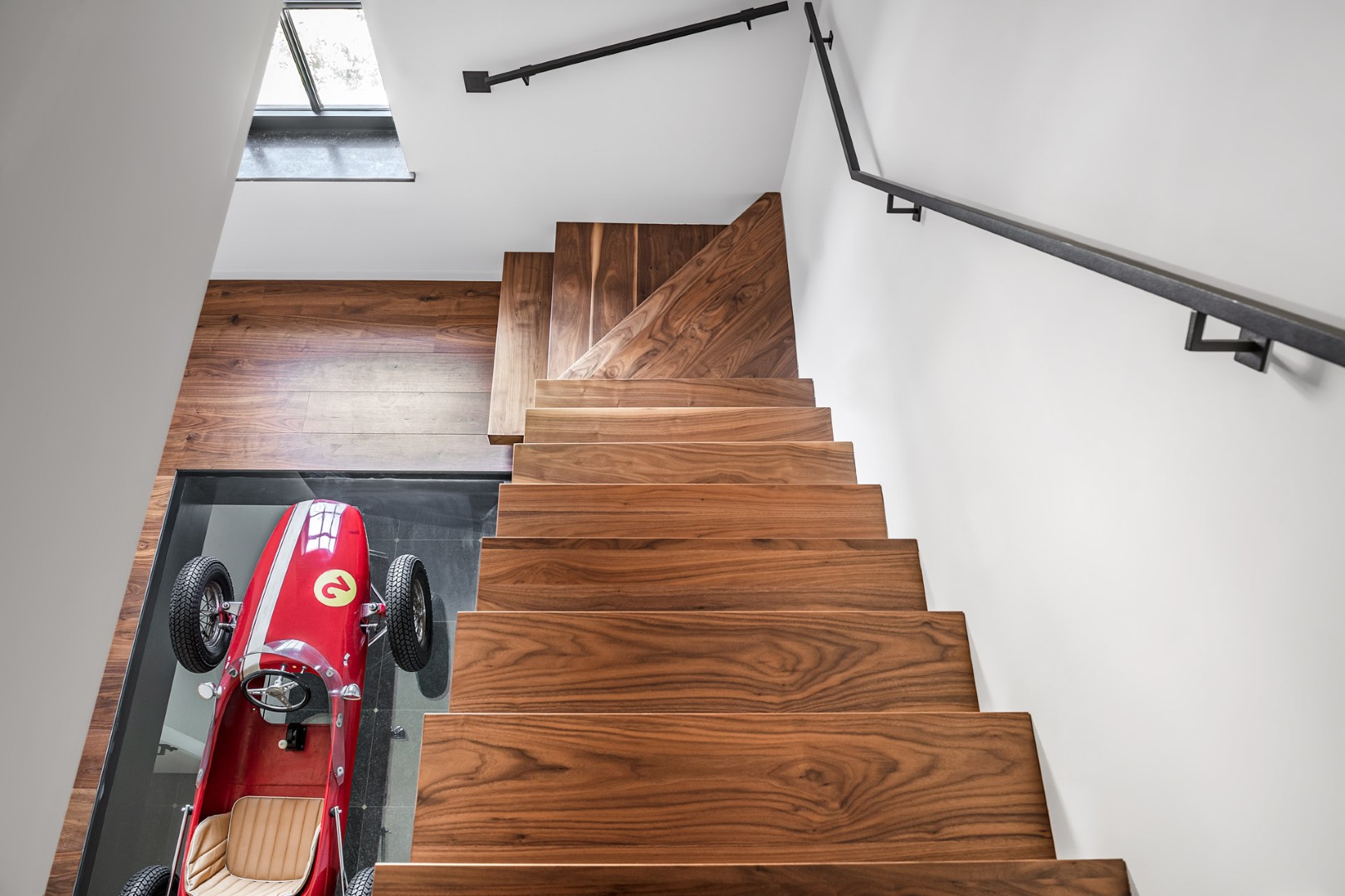 Een houten trap met zwarte leuning, met onderaan een rode speelgoedracewagen op een glazen vloer.
