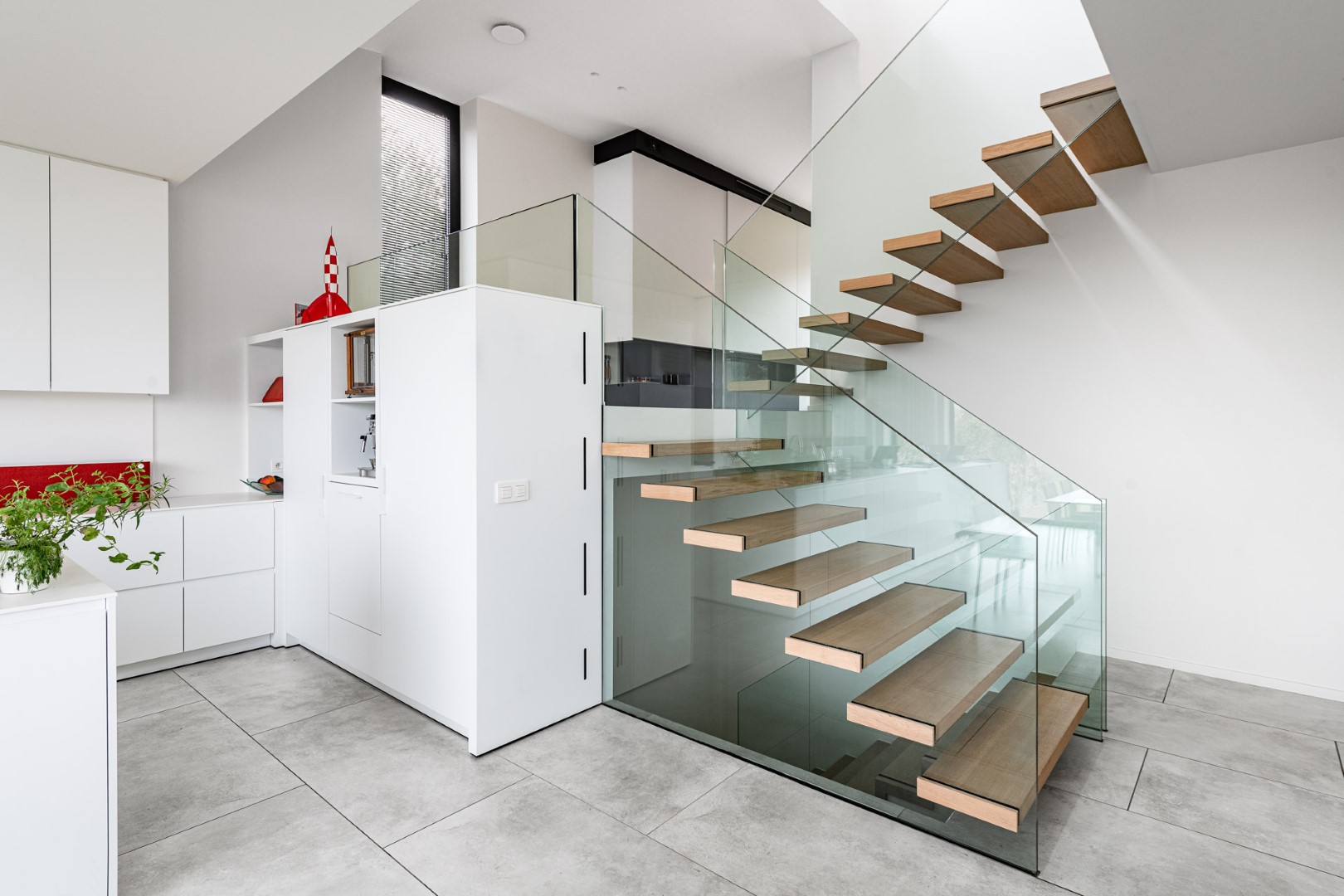 Zwevende houten trap met glazen balustrade in een strakke, witte keuken met grote grijze vloertegels en minimalistisch interieur.