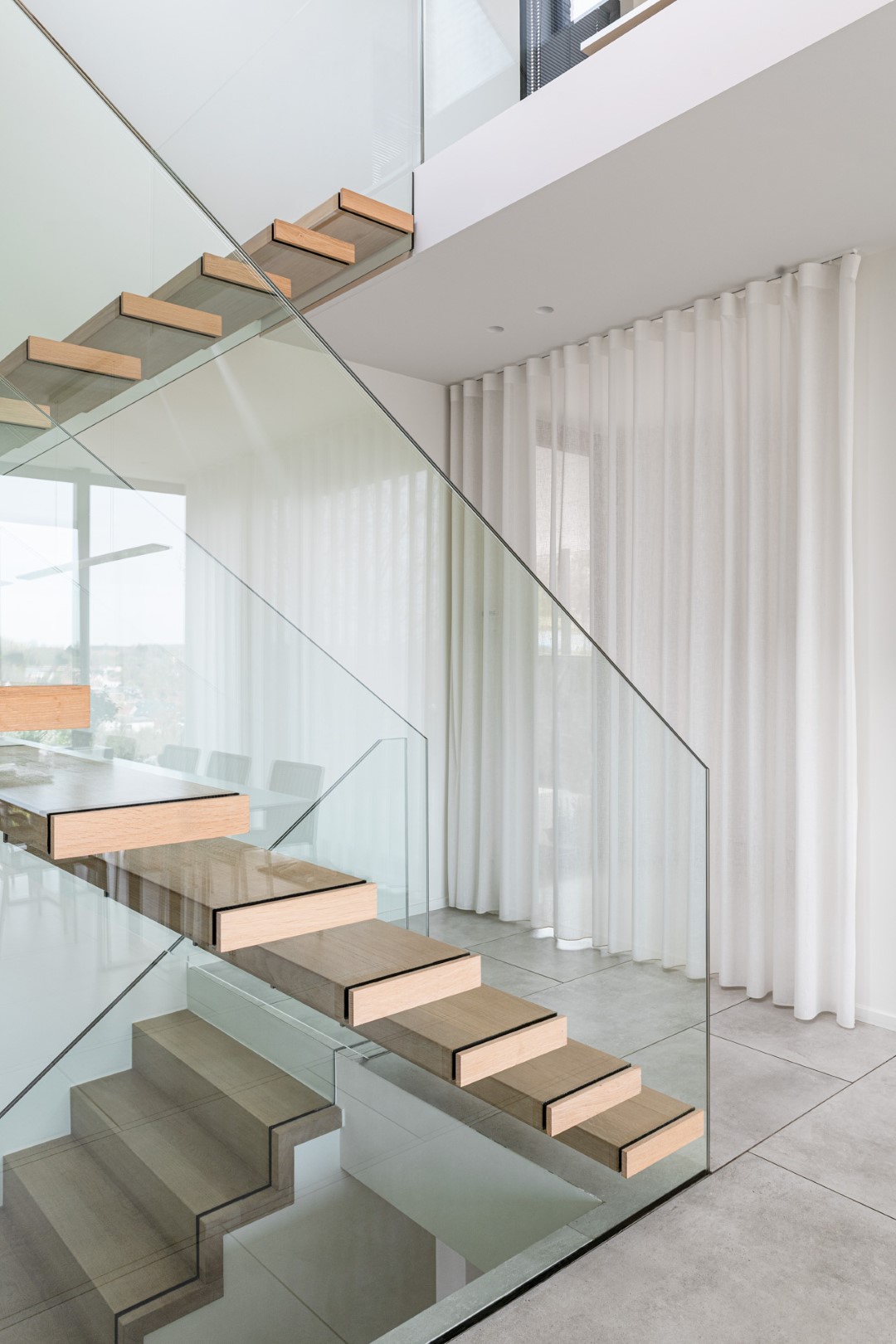 Minimalistische trap met zwevende houten treden en een strakke glazen balustrade, in een lichte moderne ruimte met grijze tegelvloer en witte gordijnen.