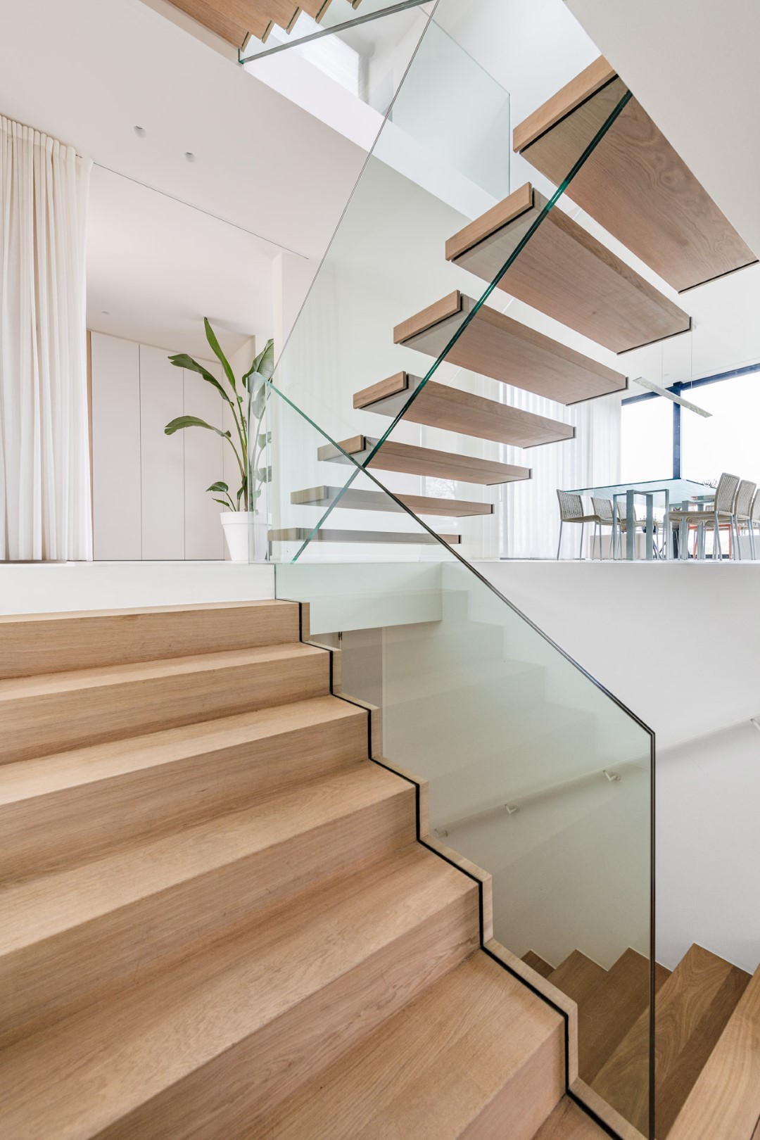 Trap met massieve houten treden en een glazen balustrade, leidend naar een verdieping met zwevende treden en een eetruimte in een helder, eigentijds interieur.