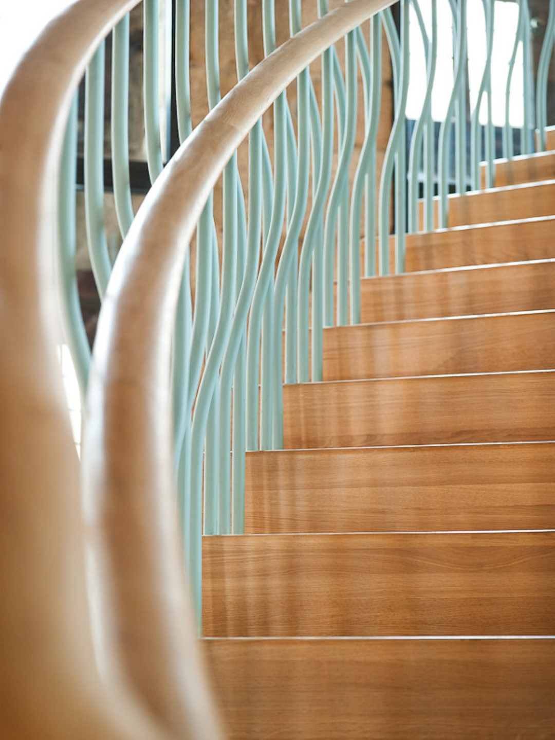 Close-up van een trap met gladde houten treden en golvende lichtblauwe metalen spijlen, omlijst door een gebogen houten leuning.