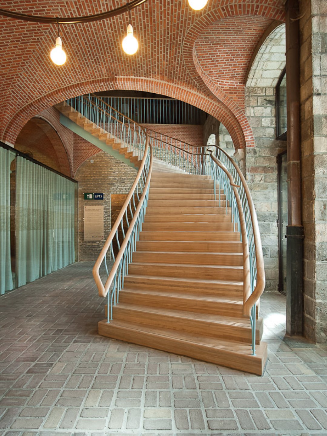 Brede houten trap met golvende metalen spijlen en houten leuning, gelegen in een historisch gebouw met bakstenen gewelven, natuurstenen muren en een vloer in visgraatpatroon.