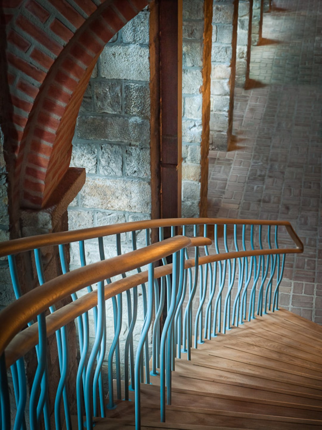 Houten trap met golvende blauwgroene spijlen en dubbele houten leuning, langs een muur van natuursteen en rode baksteenbogen in een sfeervolle, historisch ogende gang.