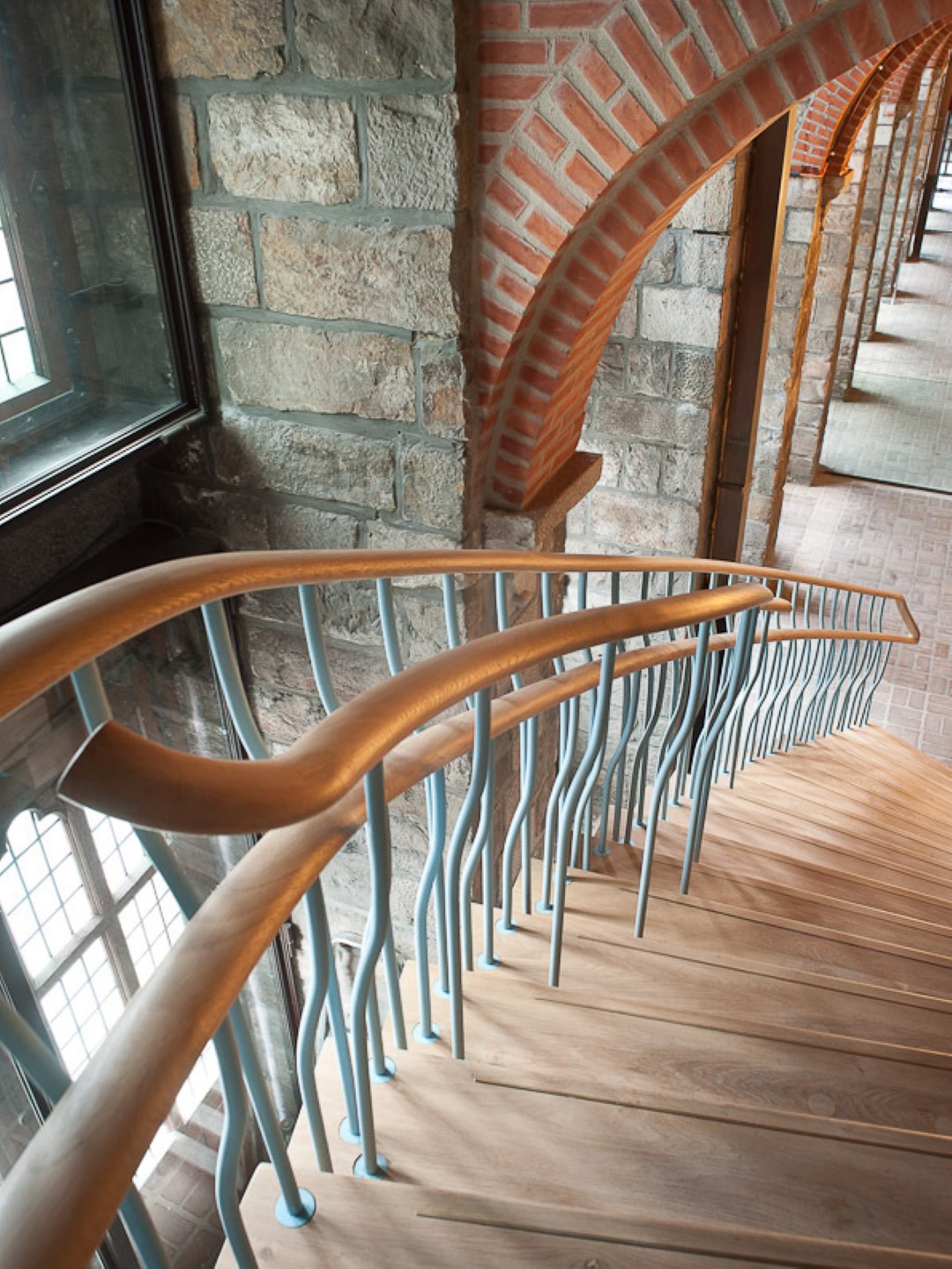 Houten trap met unieke golvende metalen spijlen en houten leuning, langs een muur van natuursteen en baksteenbogen in een historisch pand met glas-in-loodramen.