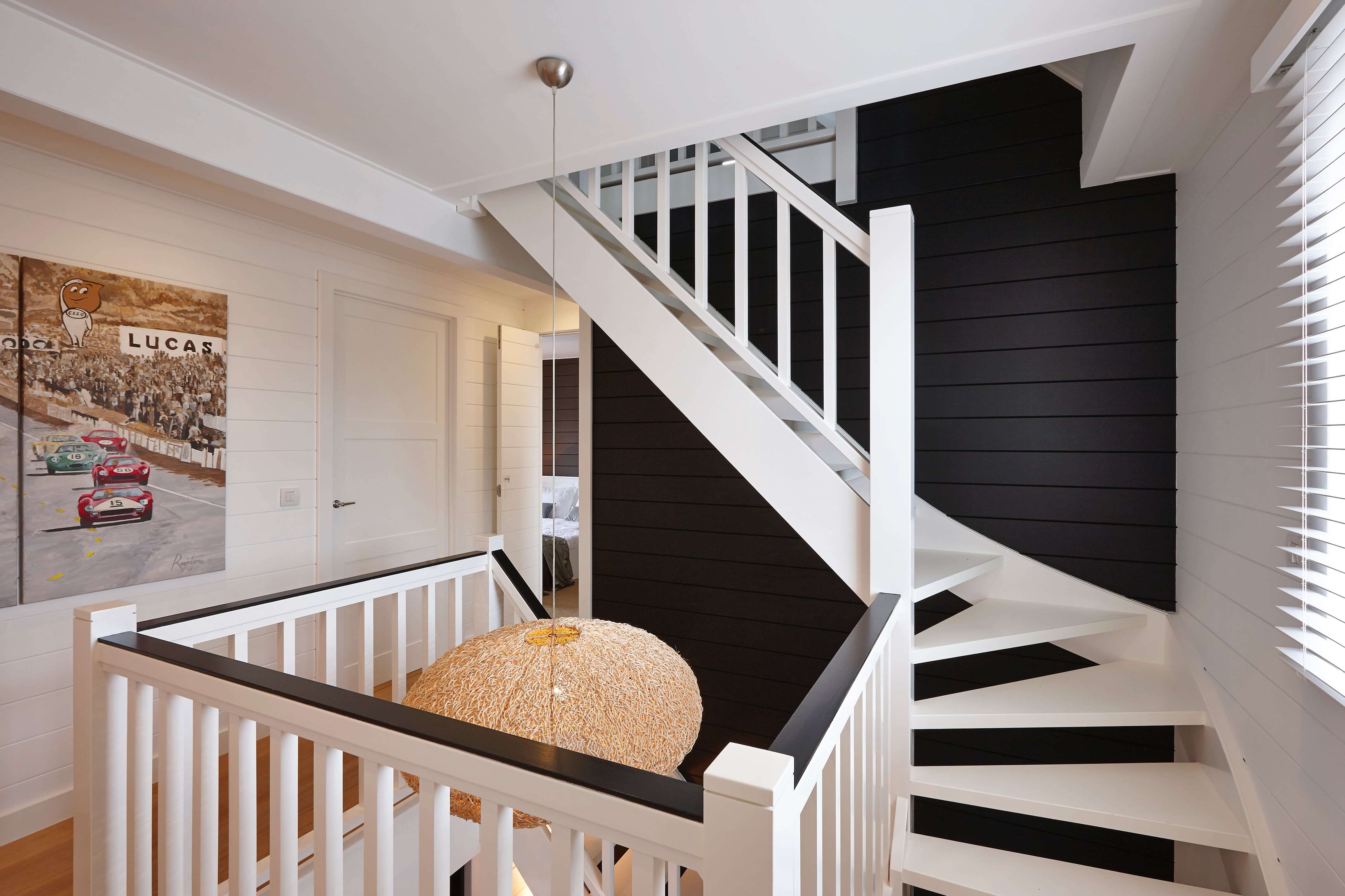 Een helder verlichte bovenverdieping met witte balustrade rondom een trapgat, een witte houten trap tegen een zwarte accentmuur, en een houten vloer die warmte toevoegt aan het verder wit afgewerkte interieur.