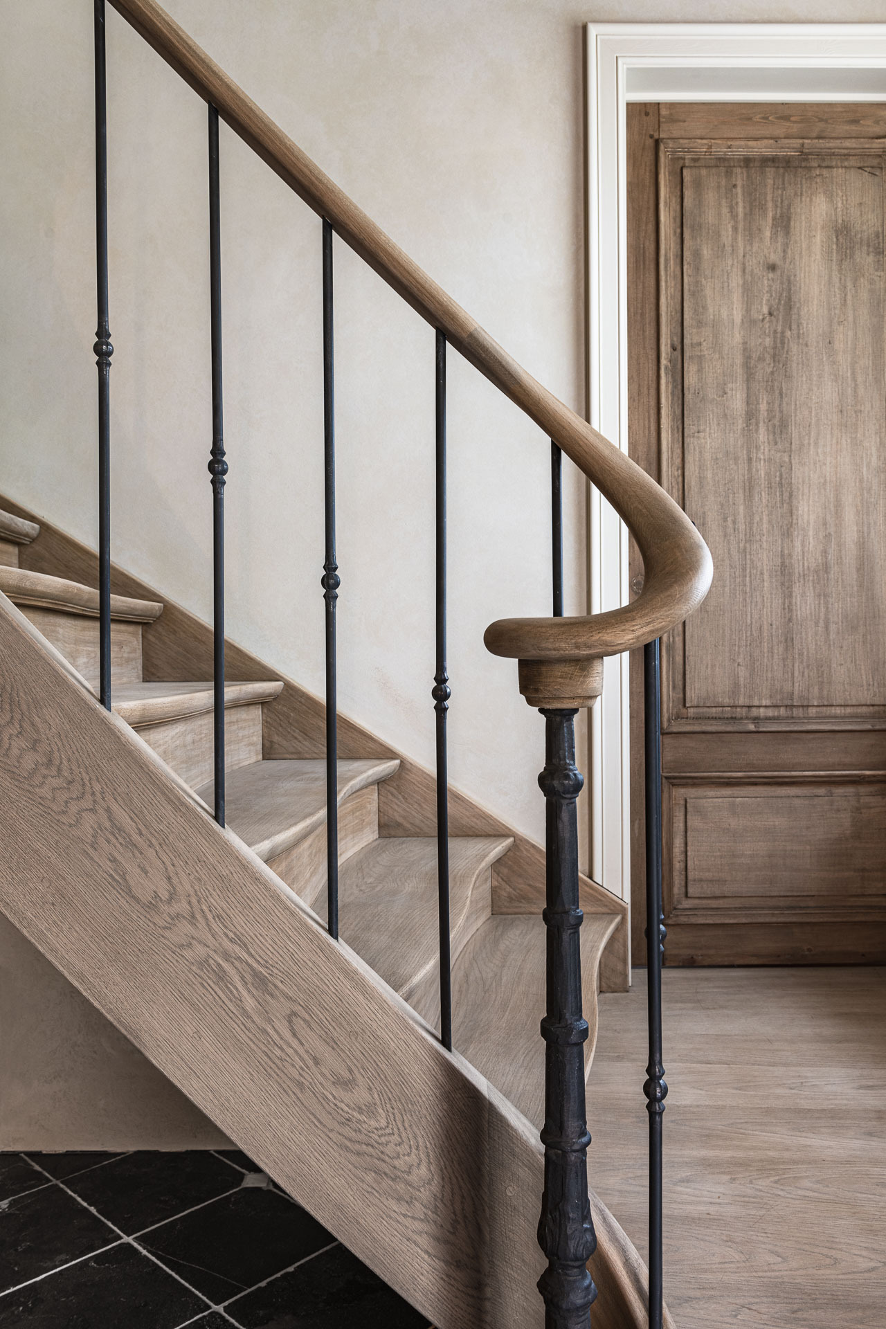 Een close-up van een houten trap met sierlijke, ronde houten leuning en verticale zwarte spijlen; op de achtergrond een rustieke houten deur en een overgang van donkere tegelvloer naar licht houten parket.