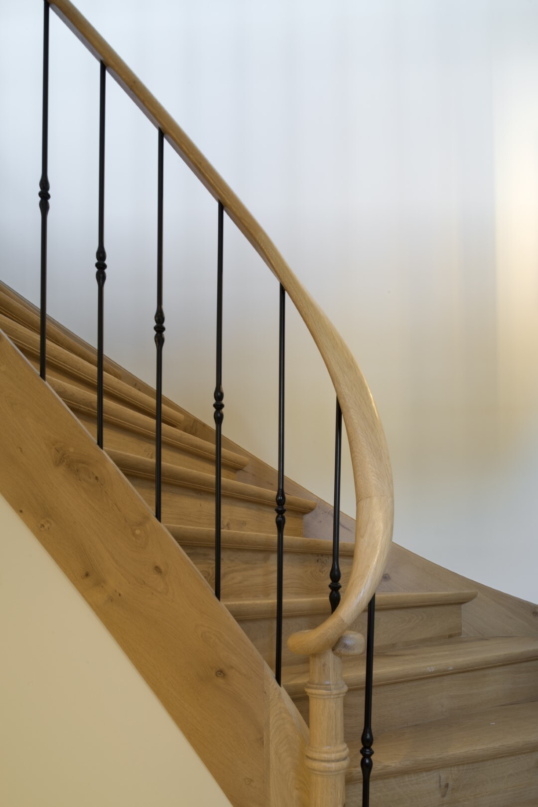 Trap met licht houten treden en afgeronde leuning, gecombineerd met fijne zwarte smeedijzeren spijlen tegen een neutrale achtergrond.