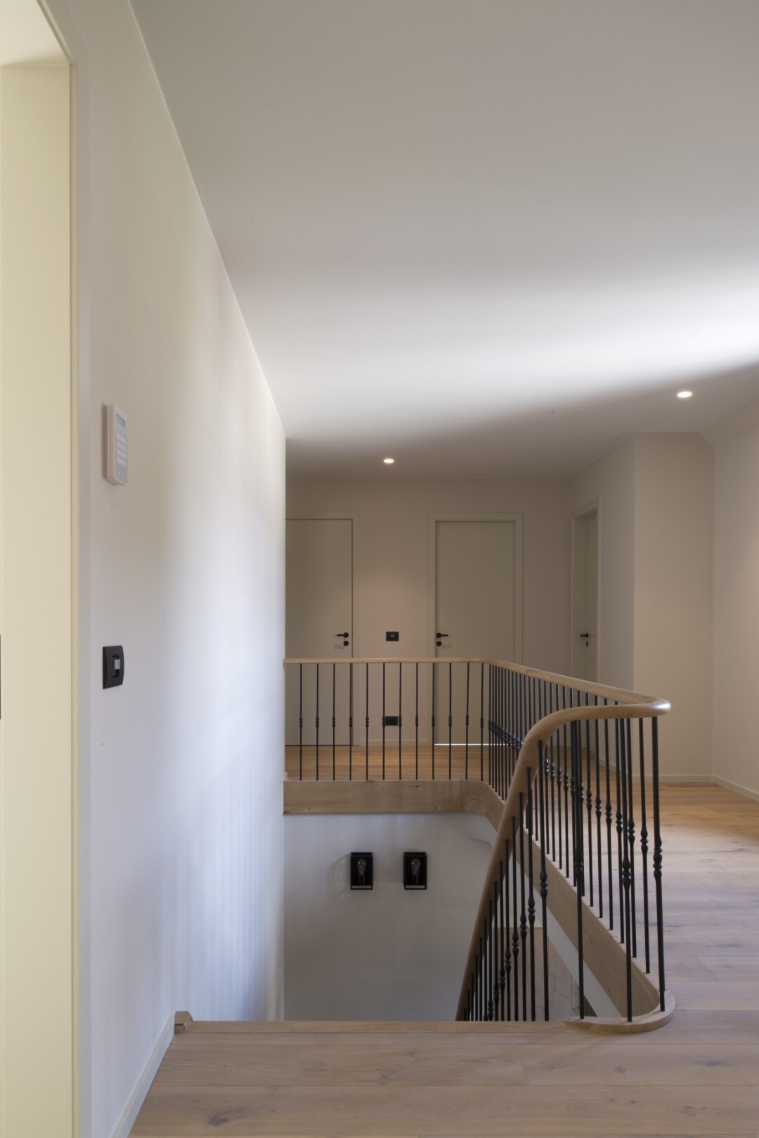 Neutrale overloop met houten vloer, trapopening met houten leuning en zwarte spijlen, en twee witte deuren aan het einde van de gang.