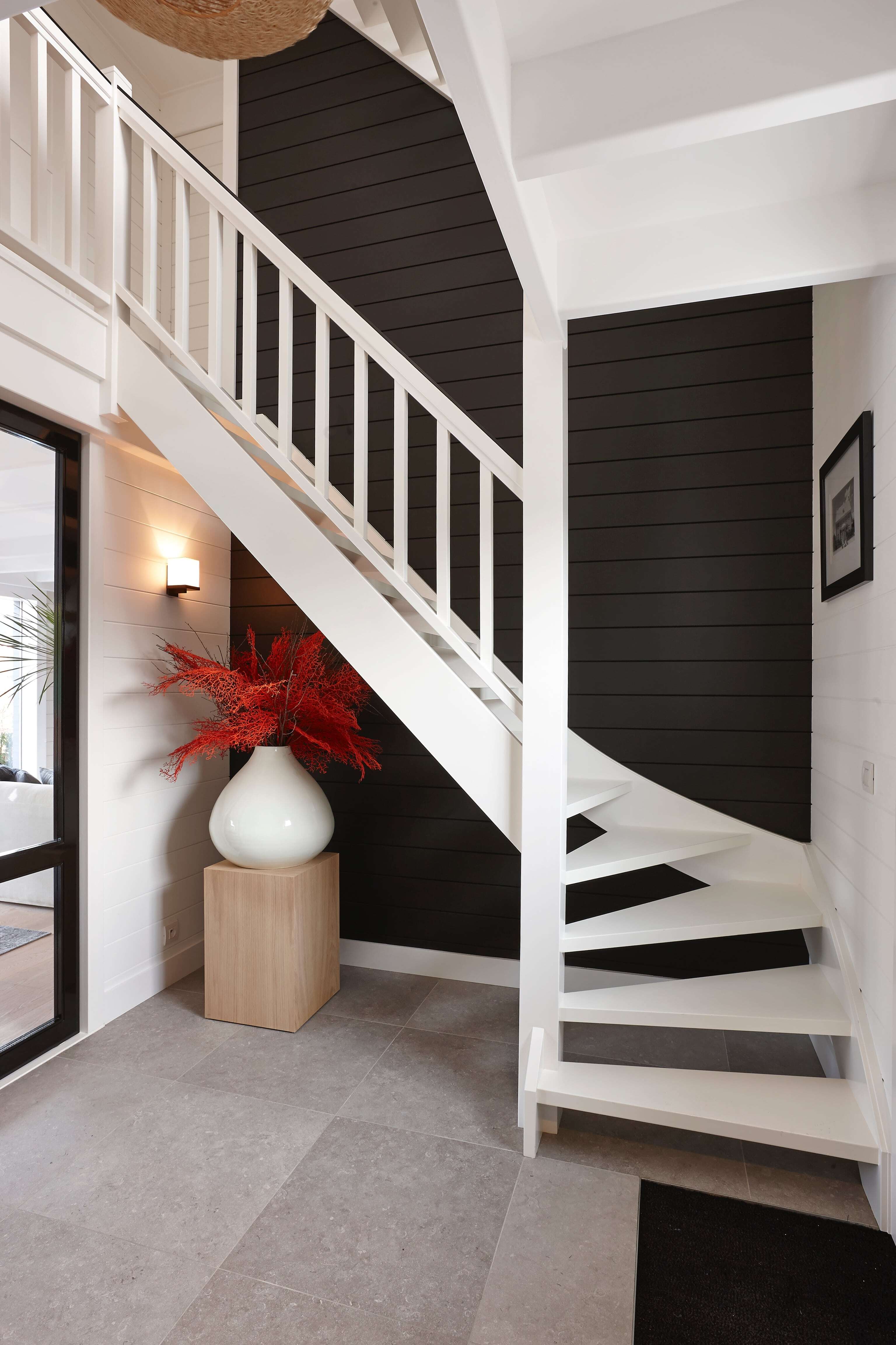 Een witte houten trap met een draai aan de onderzijde, geplaatst tegen een zwart houten lamellenmuur in een modern ingerichte hal; links staat een grote witte vaas met rode sierpluimen op een houten sokkel.