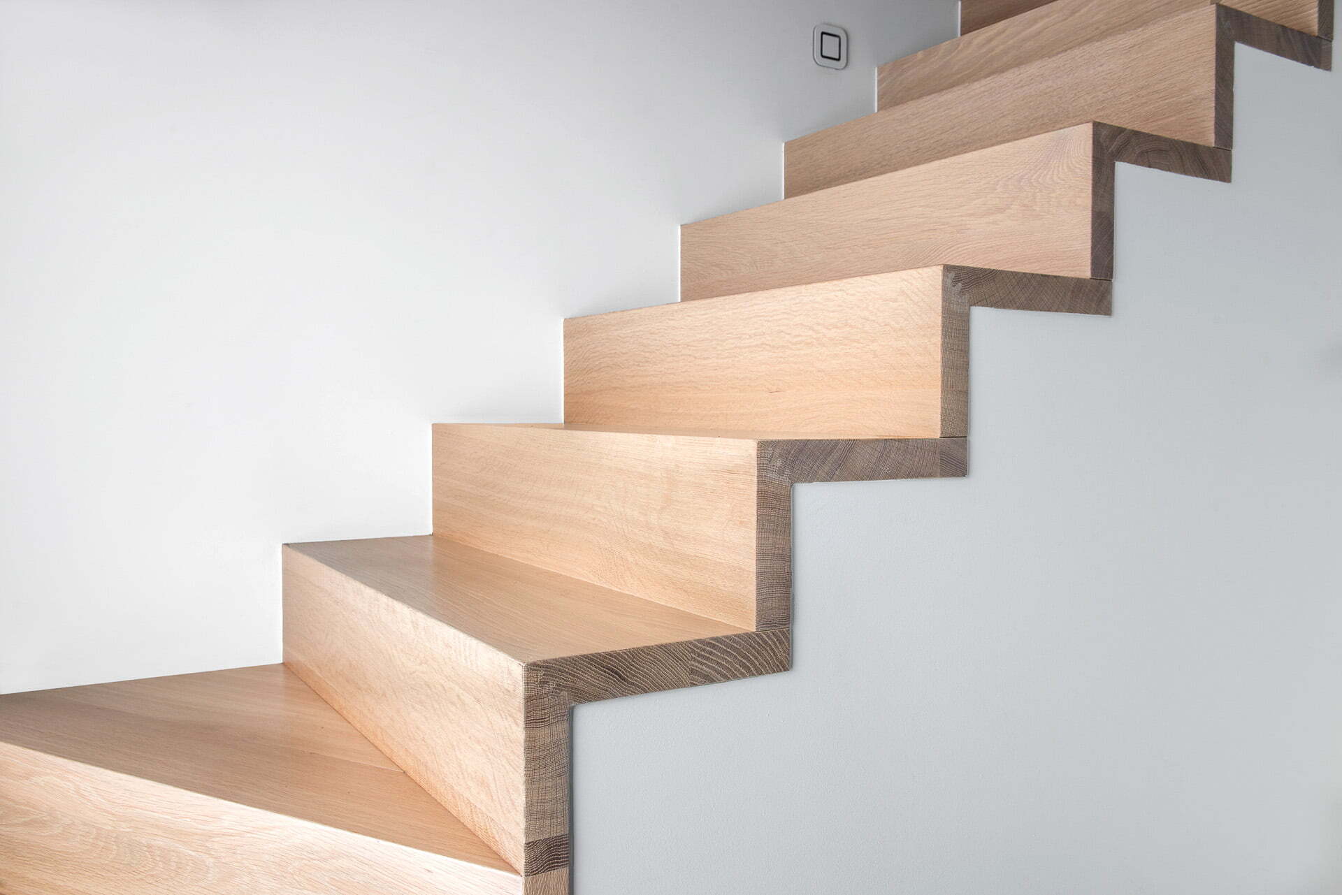 Een moderne houten trap met een opvallend zigzagprofiel langs de muur, zonder zichtbare leuning, tegen een witte achtergrond.