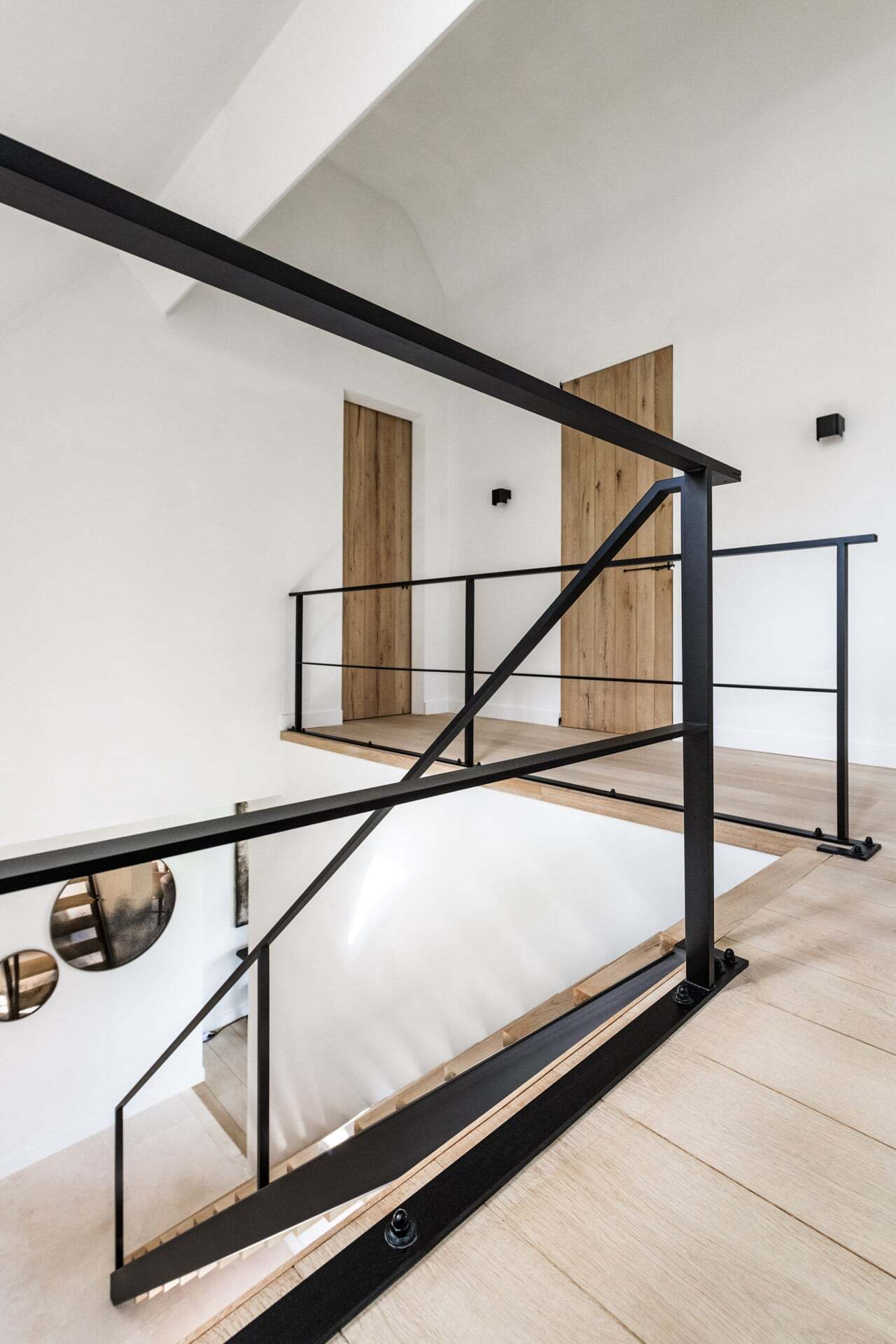 Overloop van een moderne bovenverdieping met zwarte metalen balustrade, lichte houten vloer en twee houten deuren in een strak wit interieur.