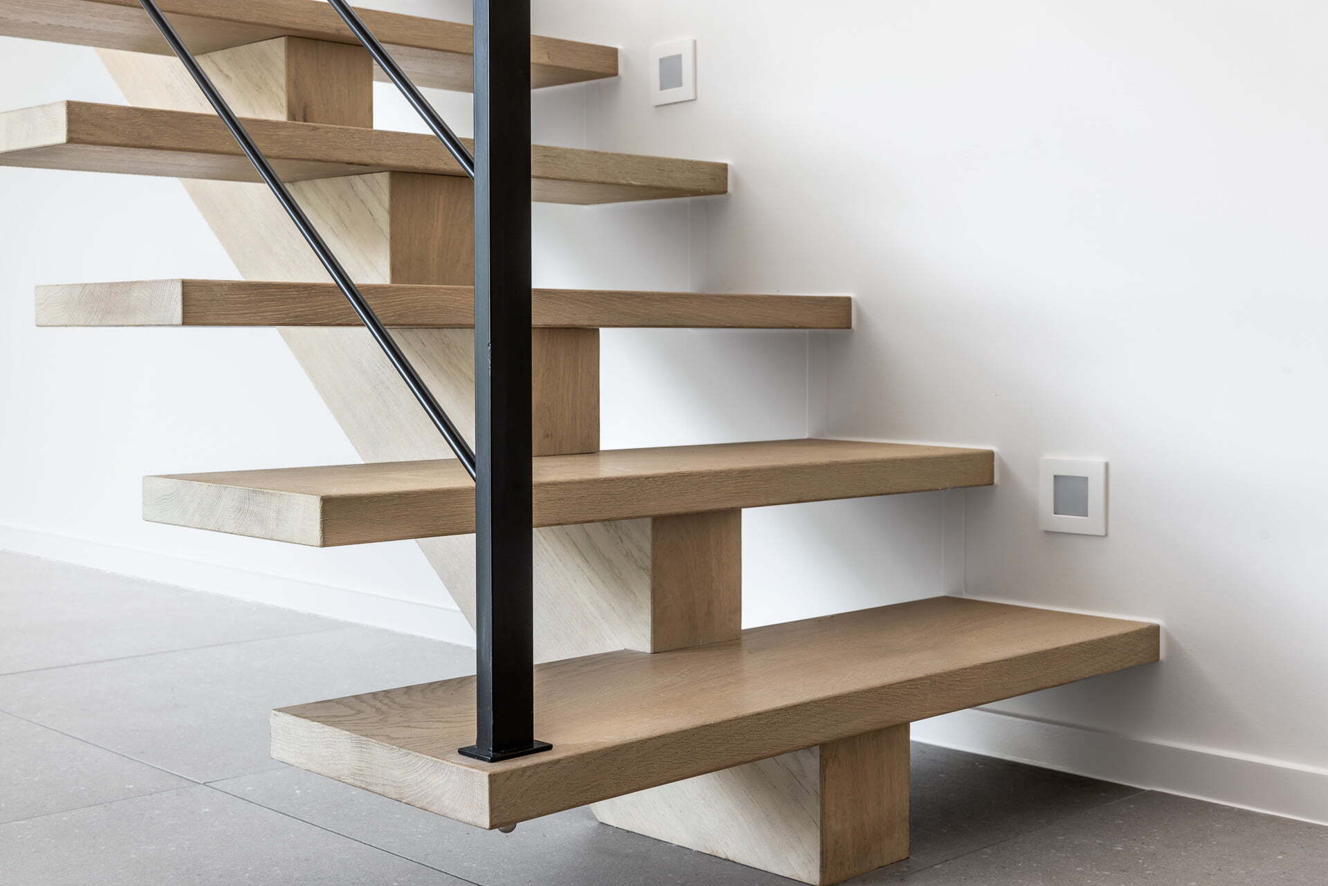Close-up van de onderzijde van een moderne open trap met dikke houten treden, zwarte metalen spijl en een witte muur met ventilatieroosters op een grijze tegelvloer.