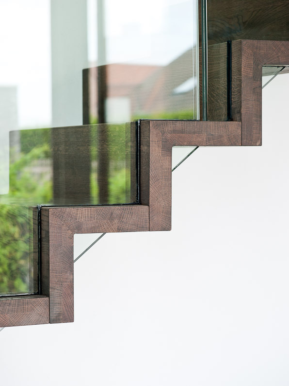 Close-up van een modern trapontwerp met donkere houten zigzagtreden en een strakke glazen balustrade, met weerspiegeling van buitenlicht en groen in het glas.