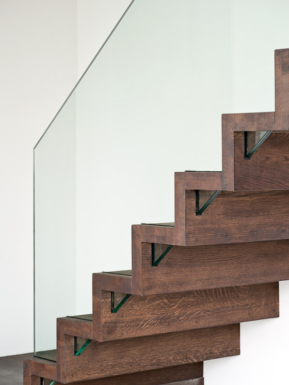 Zijaanzicht van een moderne trap met donkerbruine houten treden in zigzagvorm, ondersteund door metalen elementen en afgewerkt met een strakke glazen balustrade.