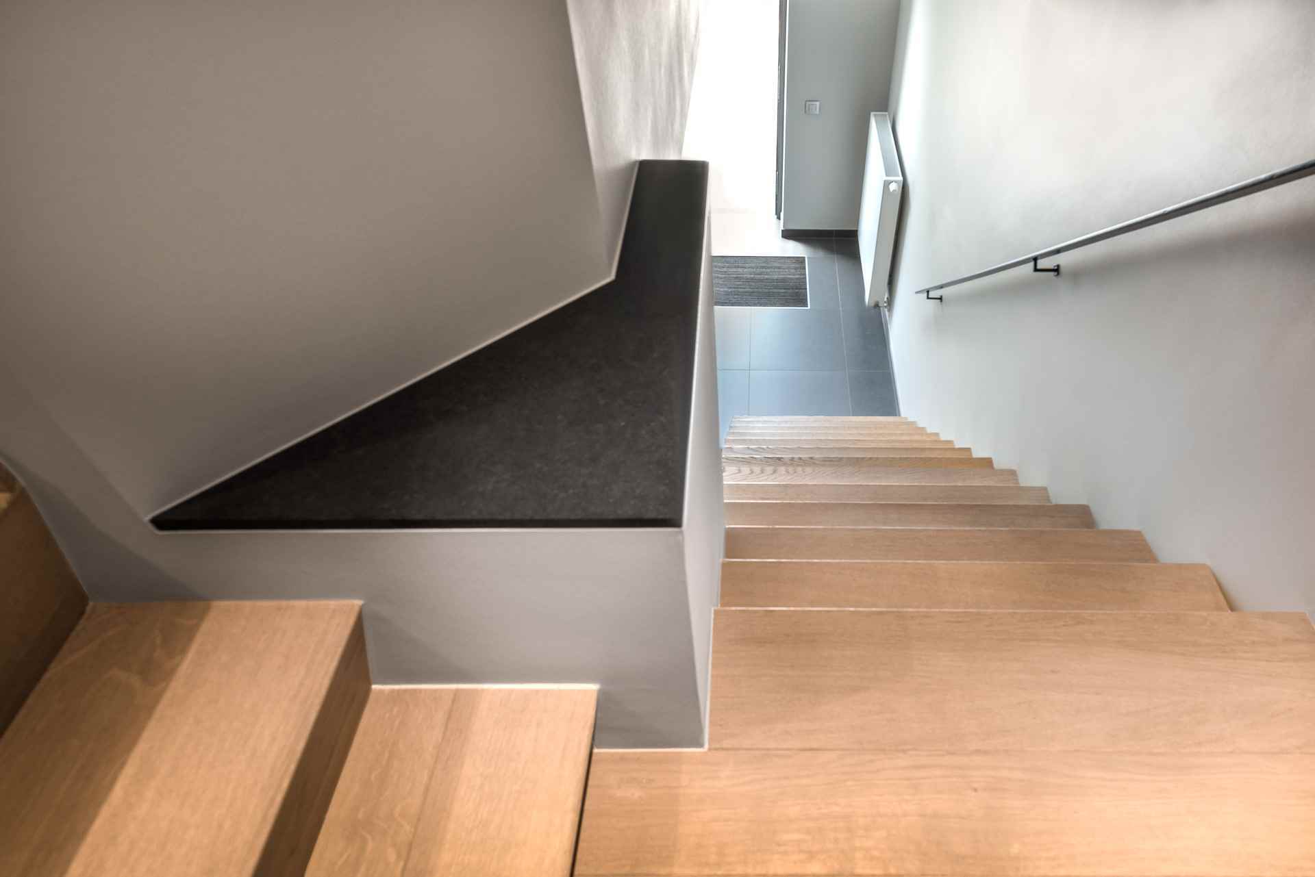 Licht houten trap met een donker tussenbordes, witte muren en een zwarte leuning, uitkomend op een hal met grijze tegelvloer.