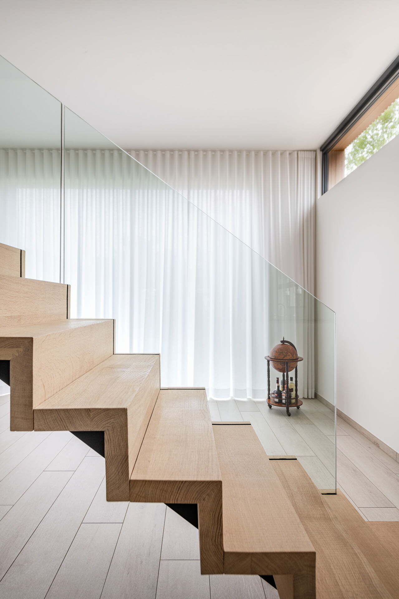 Zijaanzicht van een moderne lichte houten trap in zigzagvorm met glazen balustrade, in een ruimtelijke kamer met witte gordijnen en een decoratief houten object op een klein tafeltje.