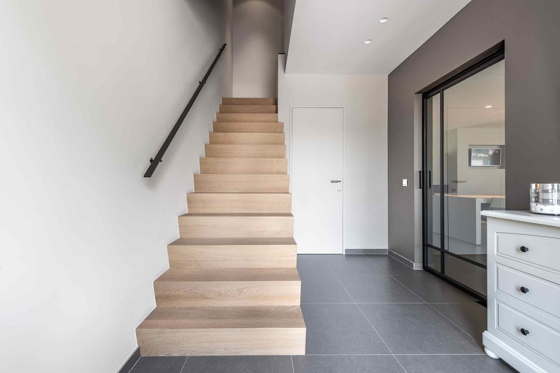 Strakke hal met een rechte houten trap, zwarte wandleuning, grote grijze vloertegels, witte muren en een glazen schuifdeur naar een andere kamer.