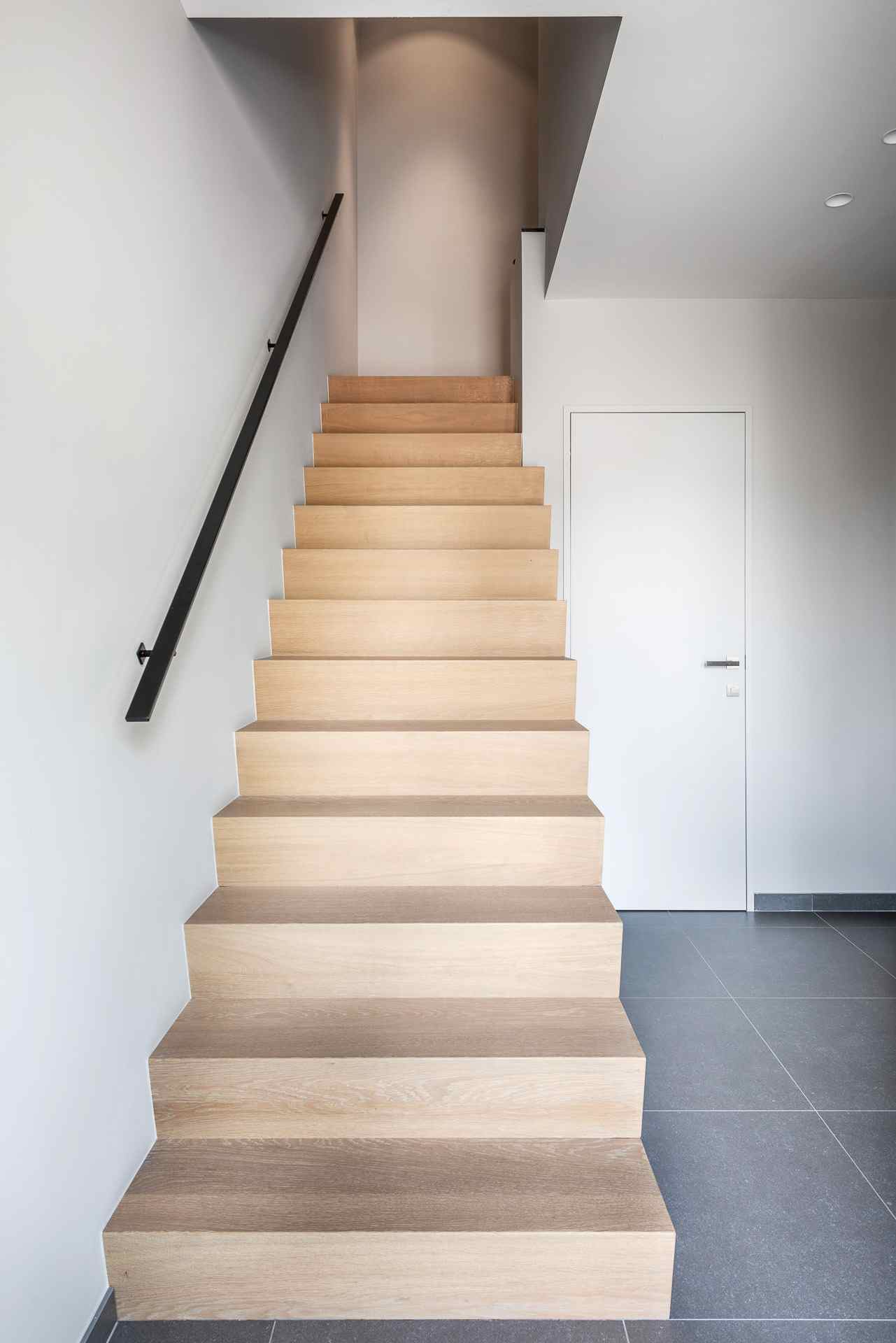 Rechte trap met lichte houten treden, zwarte wandleuning en strakke witte muren in een moderne hal met grijze tegelvloer.