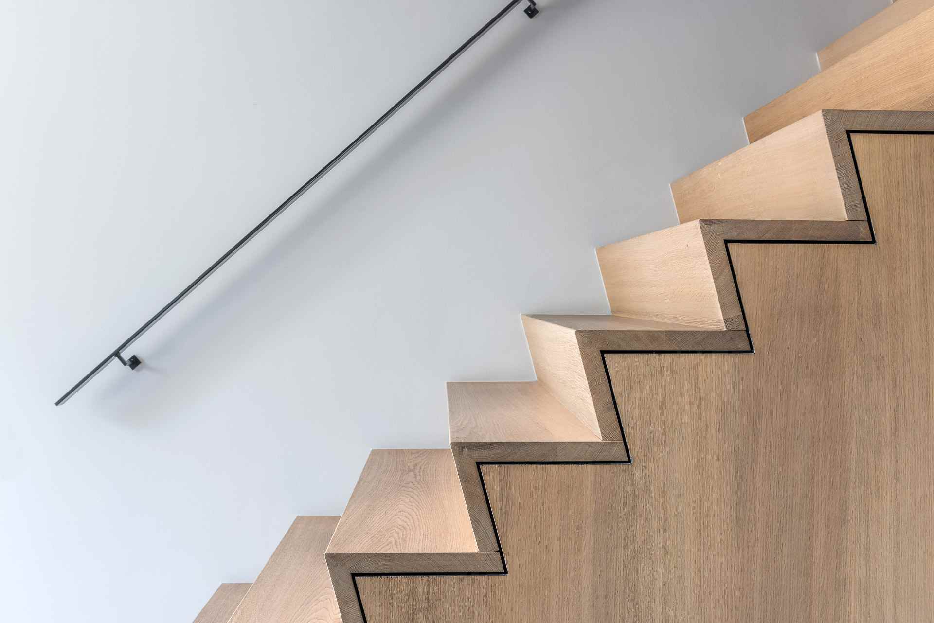 Zijaanzicht van een moderne houten trap met scherpe geometrische vormen, zwarte belijning langs de treden en een minimalistische zwarte wandleuning.