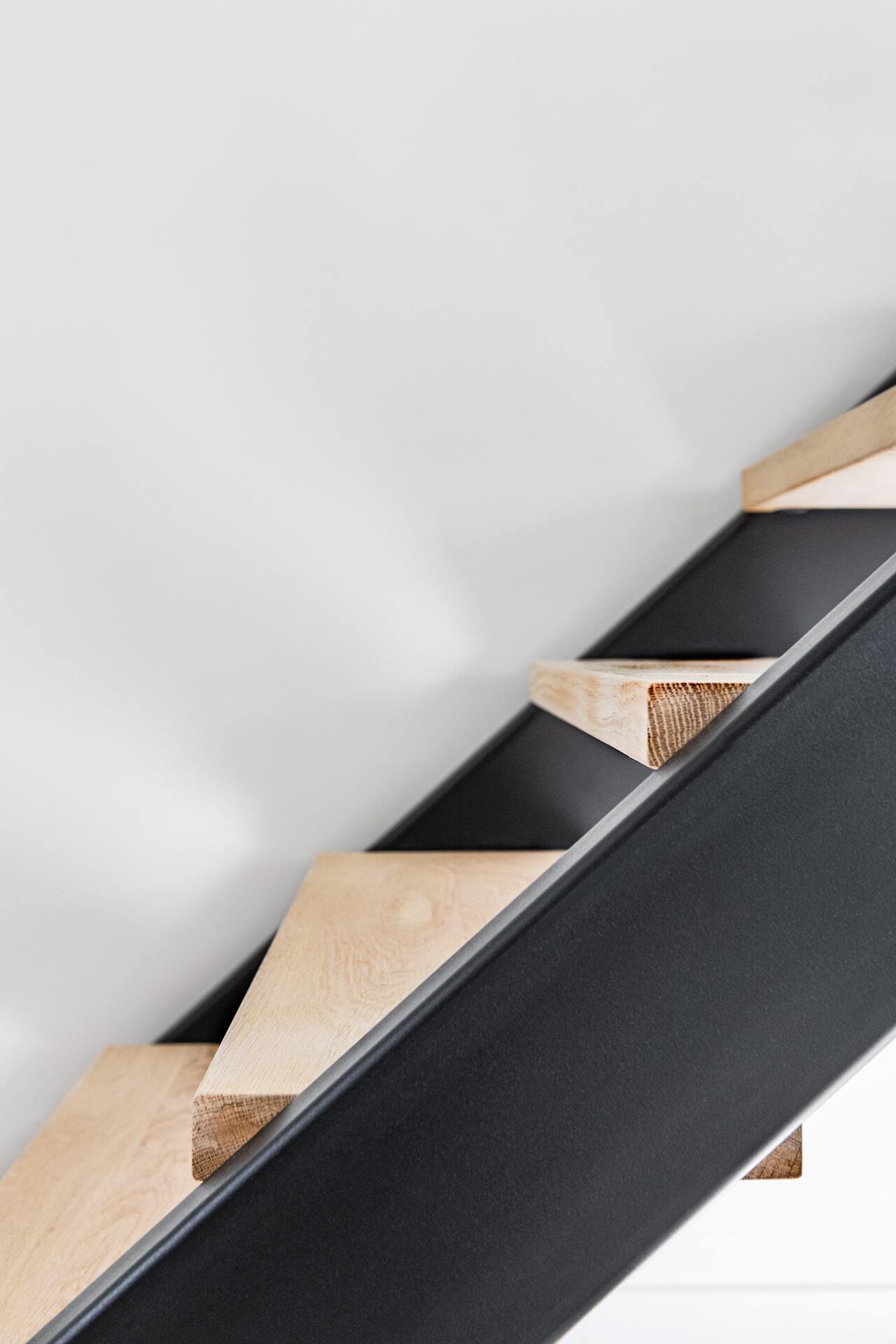 Detailopname van een moderne trap met zwevende houten treden en een zwarte stalen zijkant tegen een strakke witte muur.