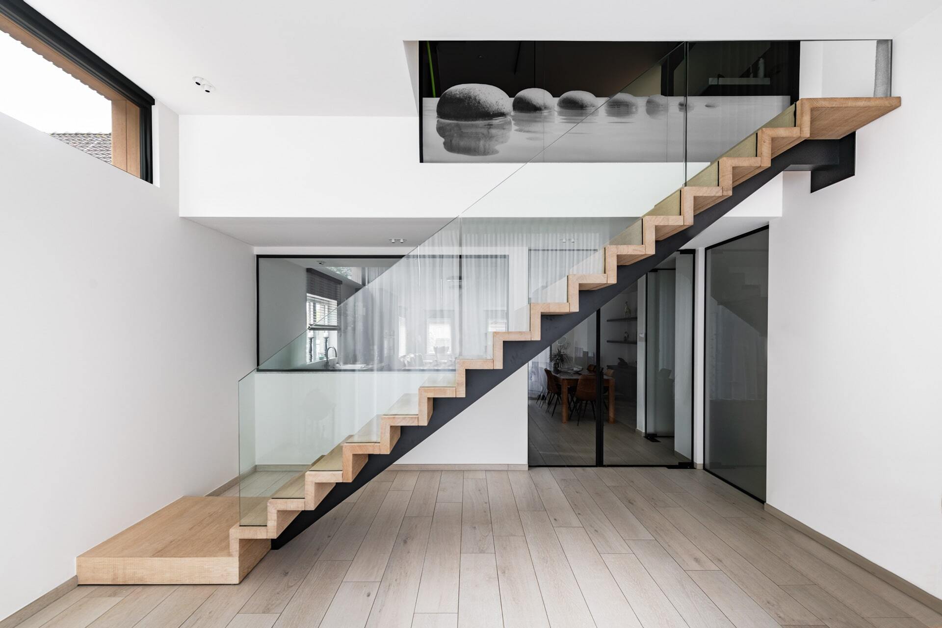 Strakke houten trap in zigzagvorm met zwarte stalen onderzijde en glazen balustrade, geplaatst in een modern interieur met lichte houten vloer en witte muren.
