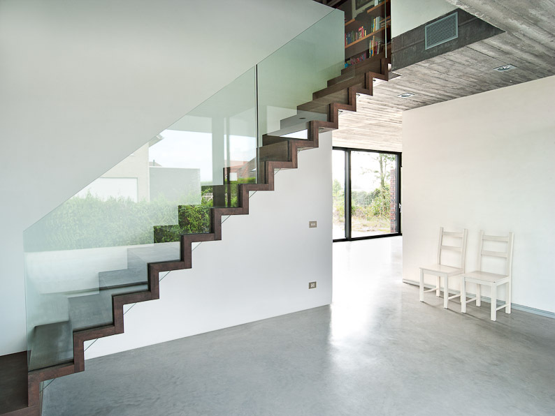 Strakke houten trap in zigzagvorm met glazen balustrade in een licht en open interieur met witte muren, betonvloer en drie witte stoelen tegen de muur.