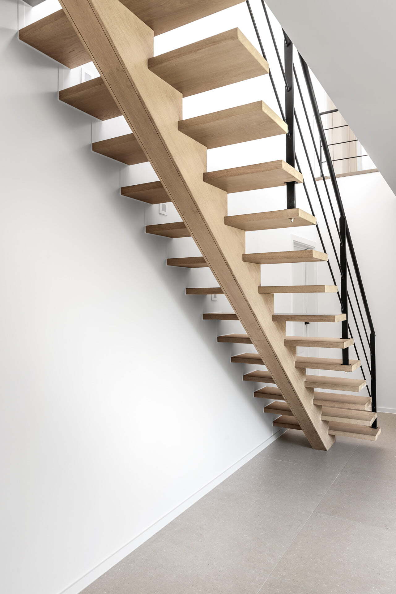 Zicht vanaf beneden op een moderne open trap met lichte houten treden en een zwarte metalen balustrade, geplaatst tegen een witte muur boven een grijze tegelvloer.