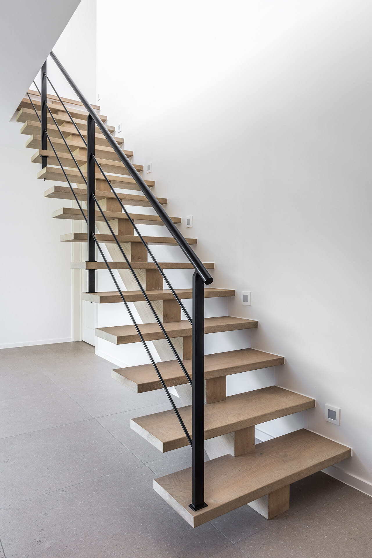 Rechte trap met dikke, open houten treden en een zwarte metalen balustrade in een minimalistisch interieur met witte muren en een grijze tegelvloer.