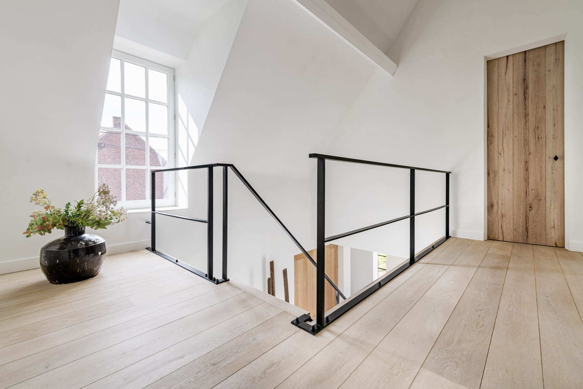 Lichte en open overloop met zwarte metalen balustrade, brede houten vloerplanken, een houten deur en een grote vaas met bloemen bij het raam.