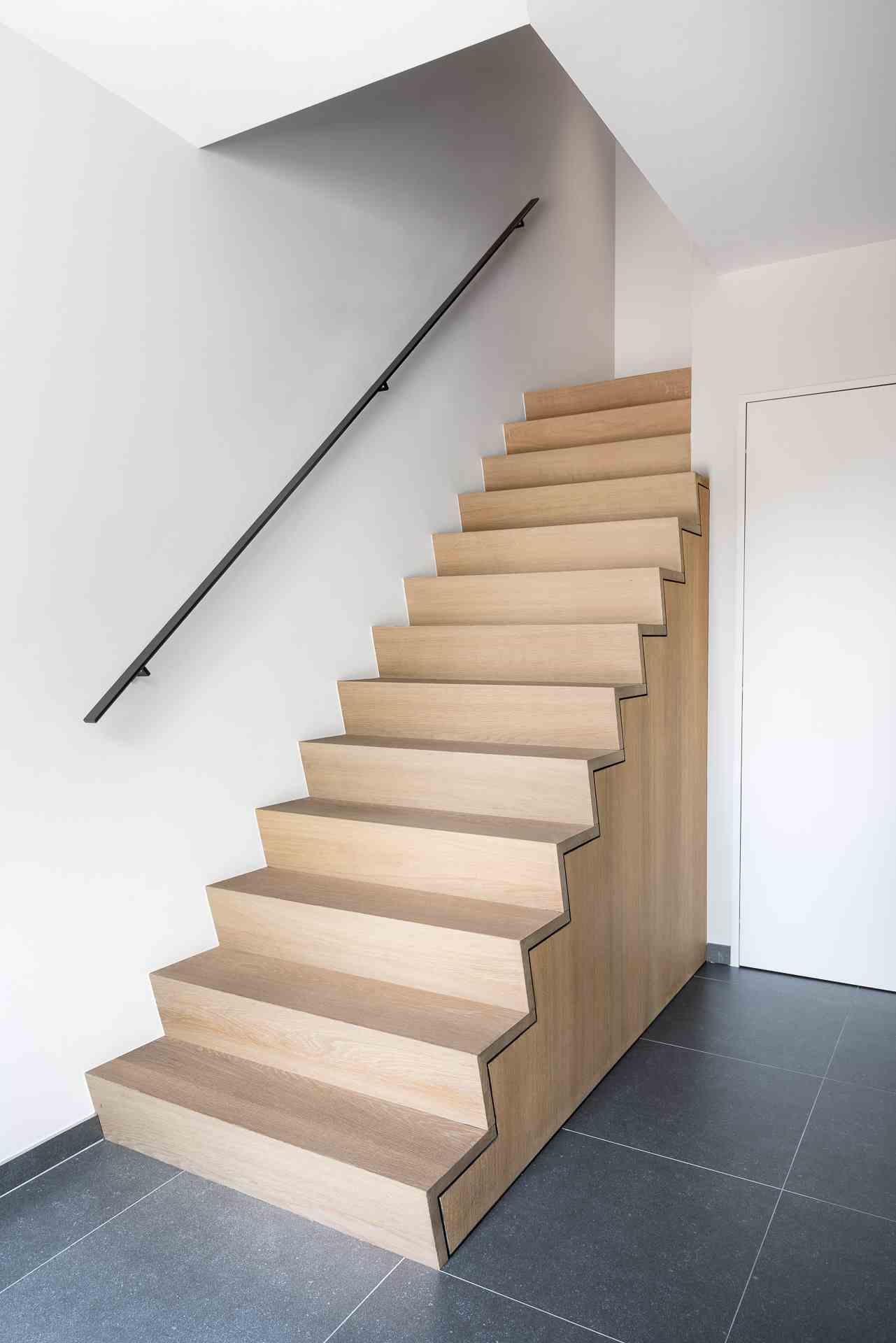 Compacte rechte trap met lichte houten treden en zijpanelen, zwarte wandleuning, witte muren en grijze tegelvloer in een strak vormgegeven hal.