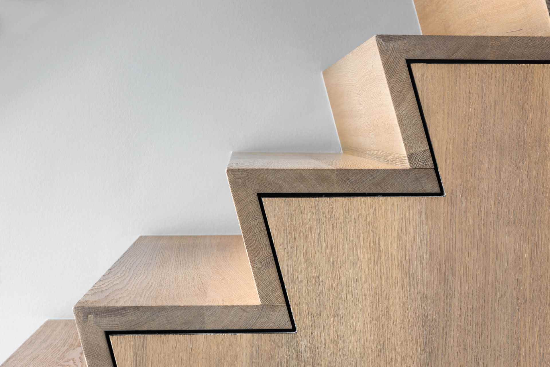 Close-up van een moderne houten trap met geometrische vormen en zwarte belijning langs de treden tegen een lichte achtergrond.