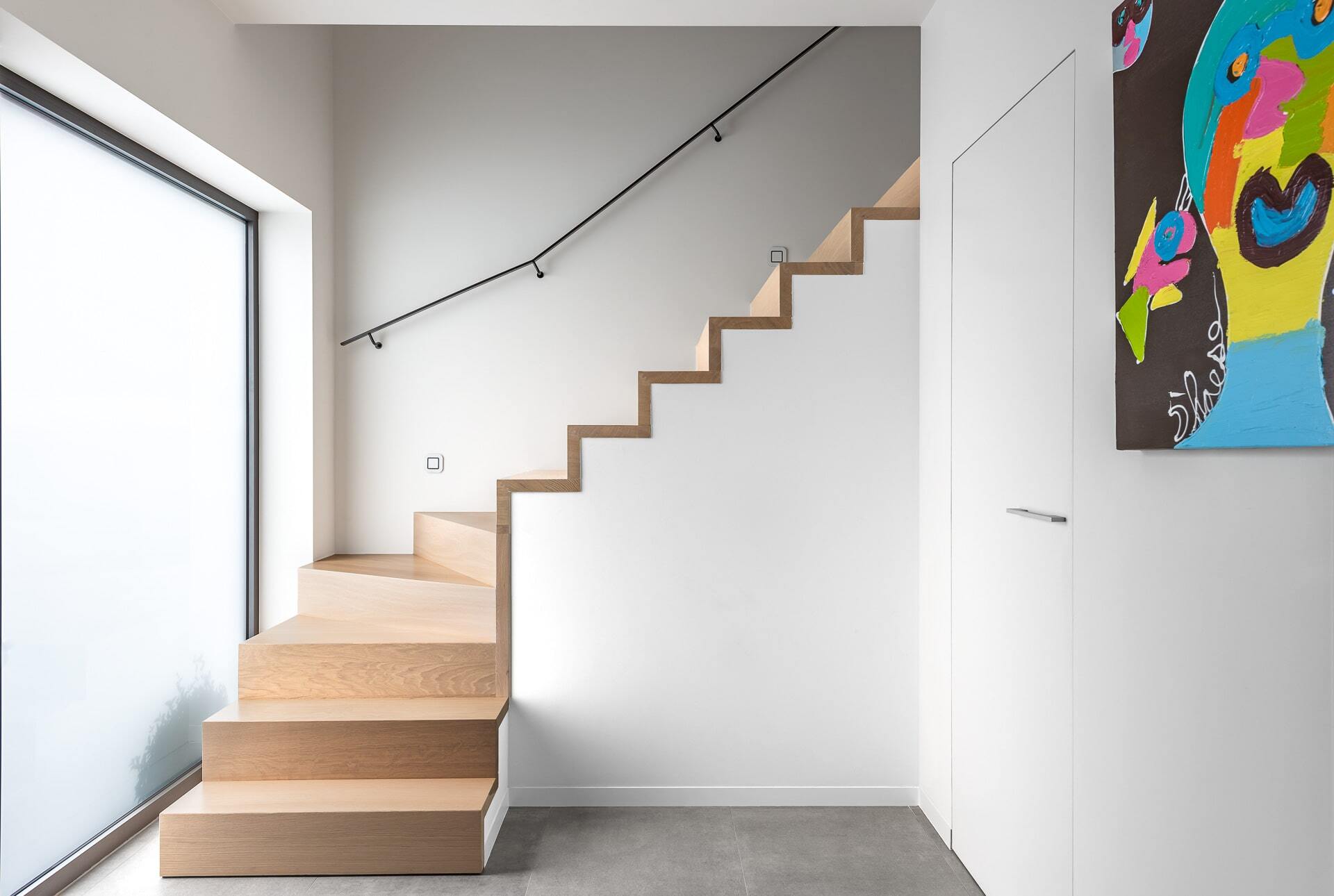 Een houten trap tegen een witte muur, met een zwarte leuning en grote raampartij links die natuurlijk licht binnenlaat; rechts hangt een felgekleurd abstract schilderij dat het minimalistische interieur contrasteert.