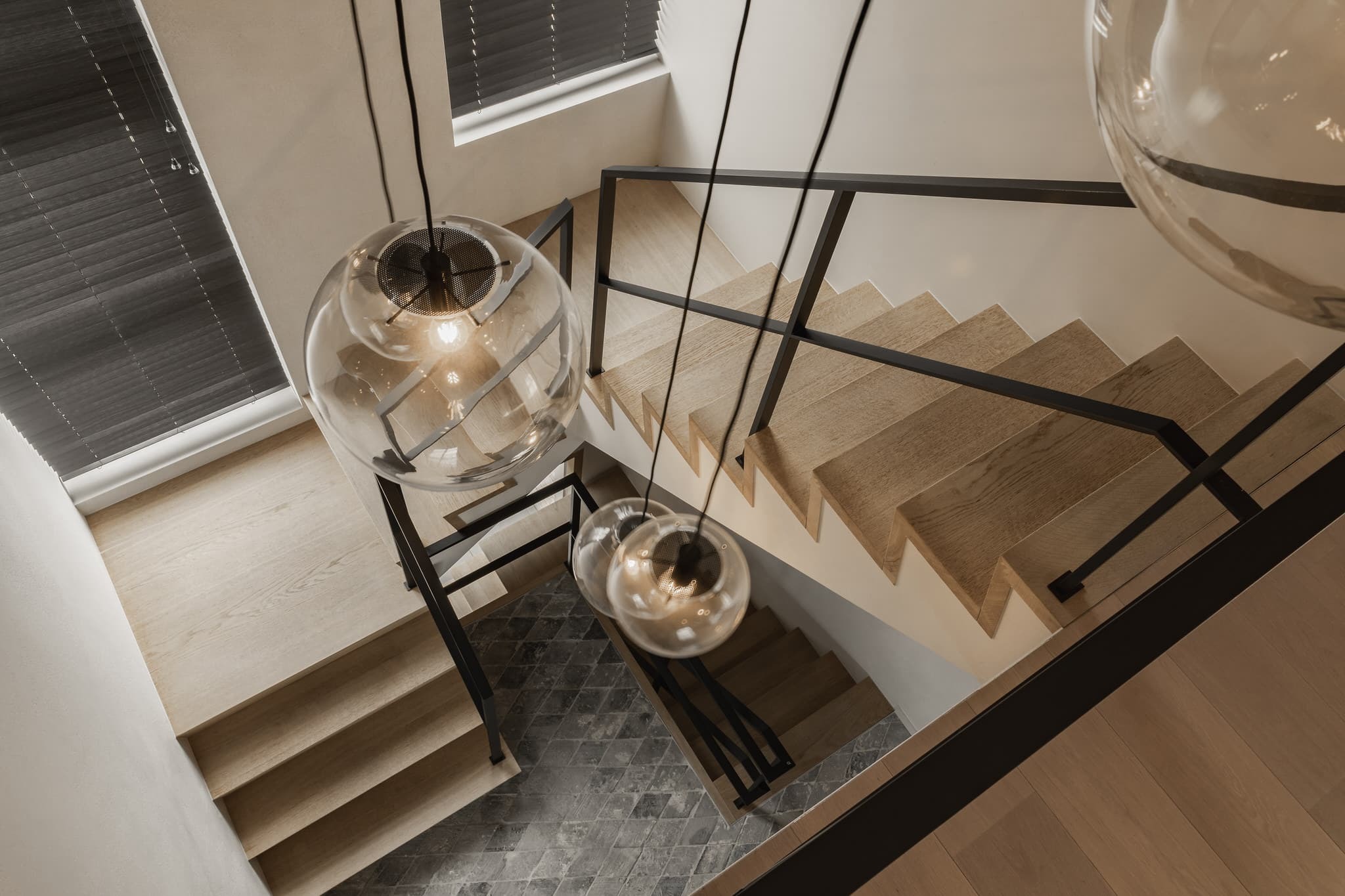 Meerdere trapdelen met lichte houten treden en zwarte metalen leuningen in een stijlvolle vide, verlicht door ronde glazen hanglampen en omringd door ramen met zwarte jaloezieën.