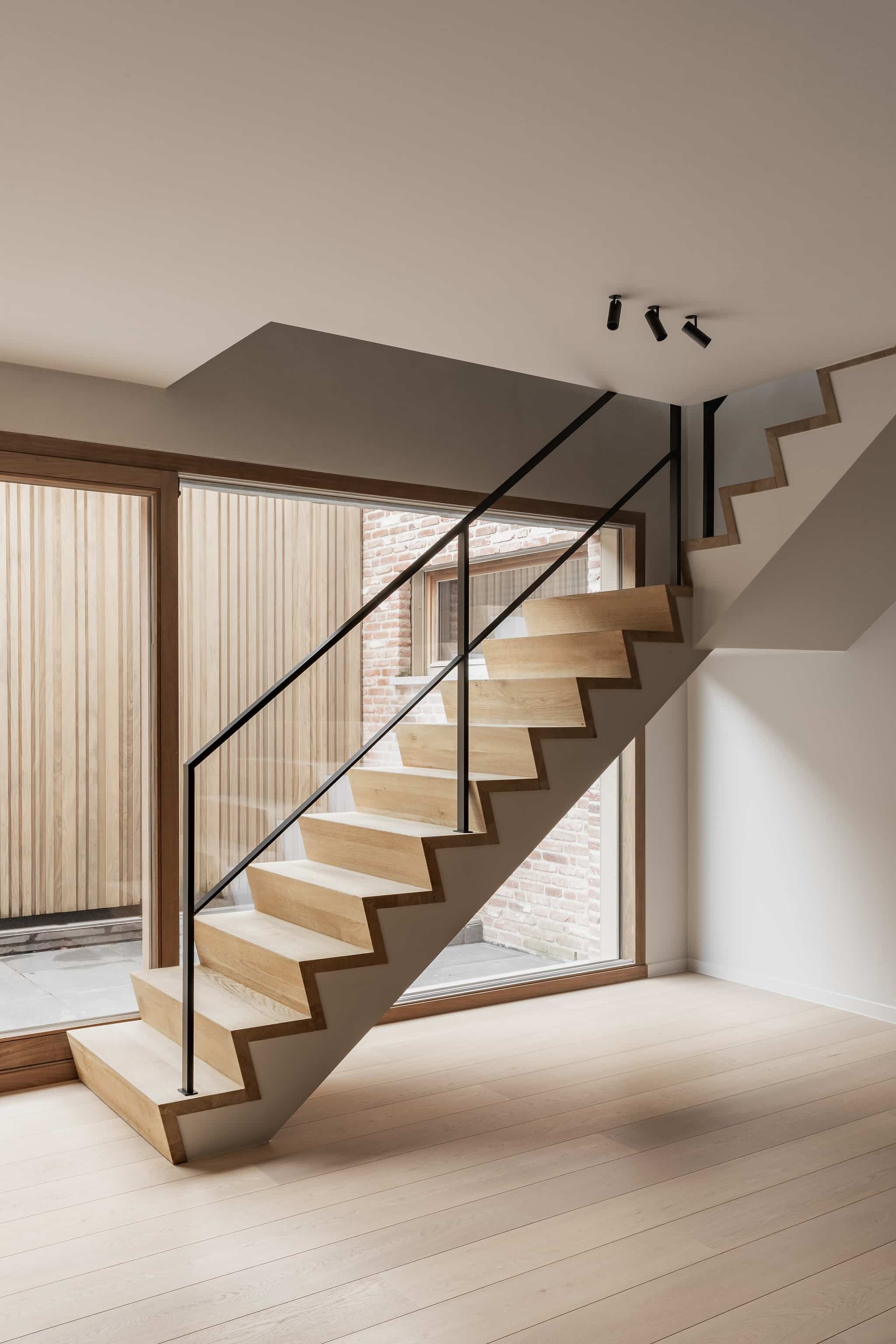 Moderne trap met lichte houten treden en een zwarte metalen leuning, in een minimalistisch interieur met houten vloer en grote ramen.