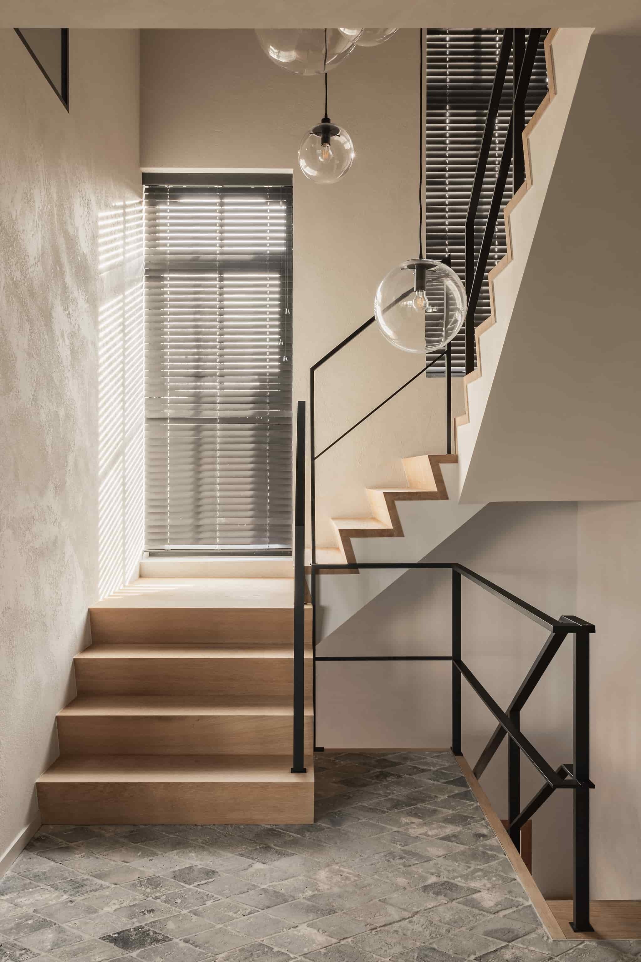 Lichte trapopgang met houten treden, zwarte metalen leuning, ronde glazen hanglampen en een groot raam met zwarte jaloezieën boven een vloer met grijstinten.