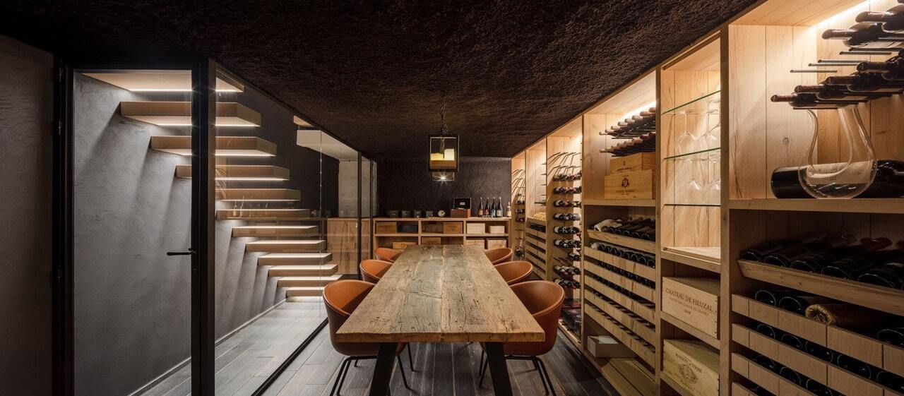 Sfeervolle wijnkelder met houten rekken en een lange eettafel, grenzend aan een moderne trap met zwevende treden en geïntegreerde ledverlichting achter glazen wand.