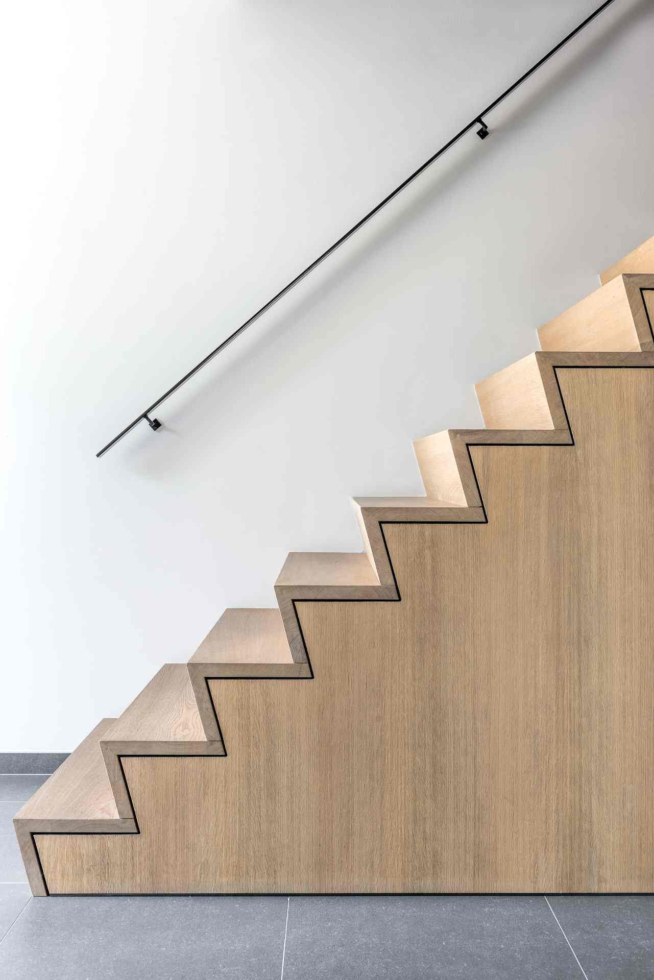 Zijaanzicht van een moderne trap met zigzagvormige houten bekleding, zwarte belijning en een slanke zwarte wandleuning tegen een witte muur en grijze tegelvloer.