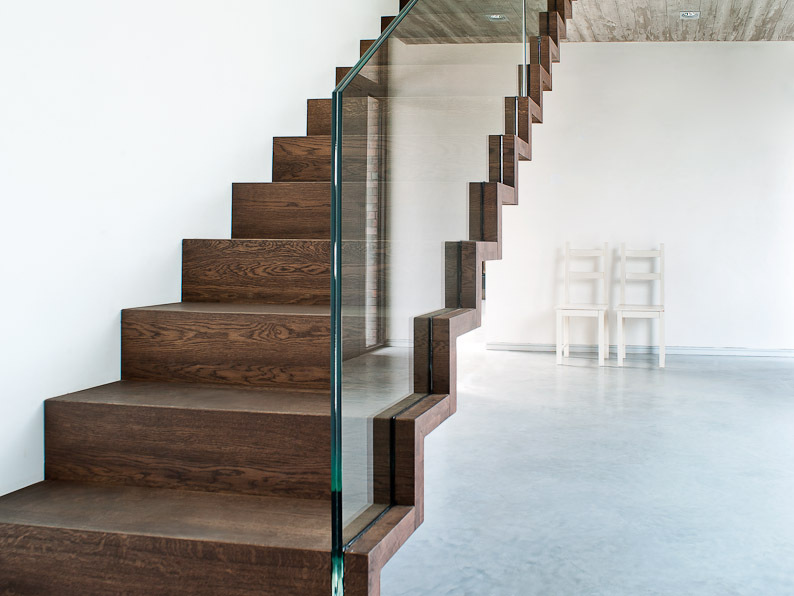 Moderne trap in zigzagvorm met donkere houten treden en een glazen balustrade, geplaatst in een minimalistische ruimte met betonvloer en witte muren.
