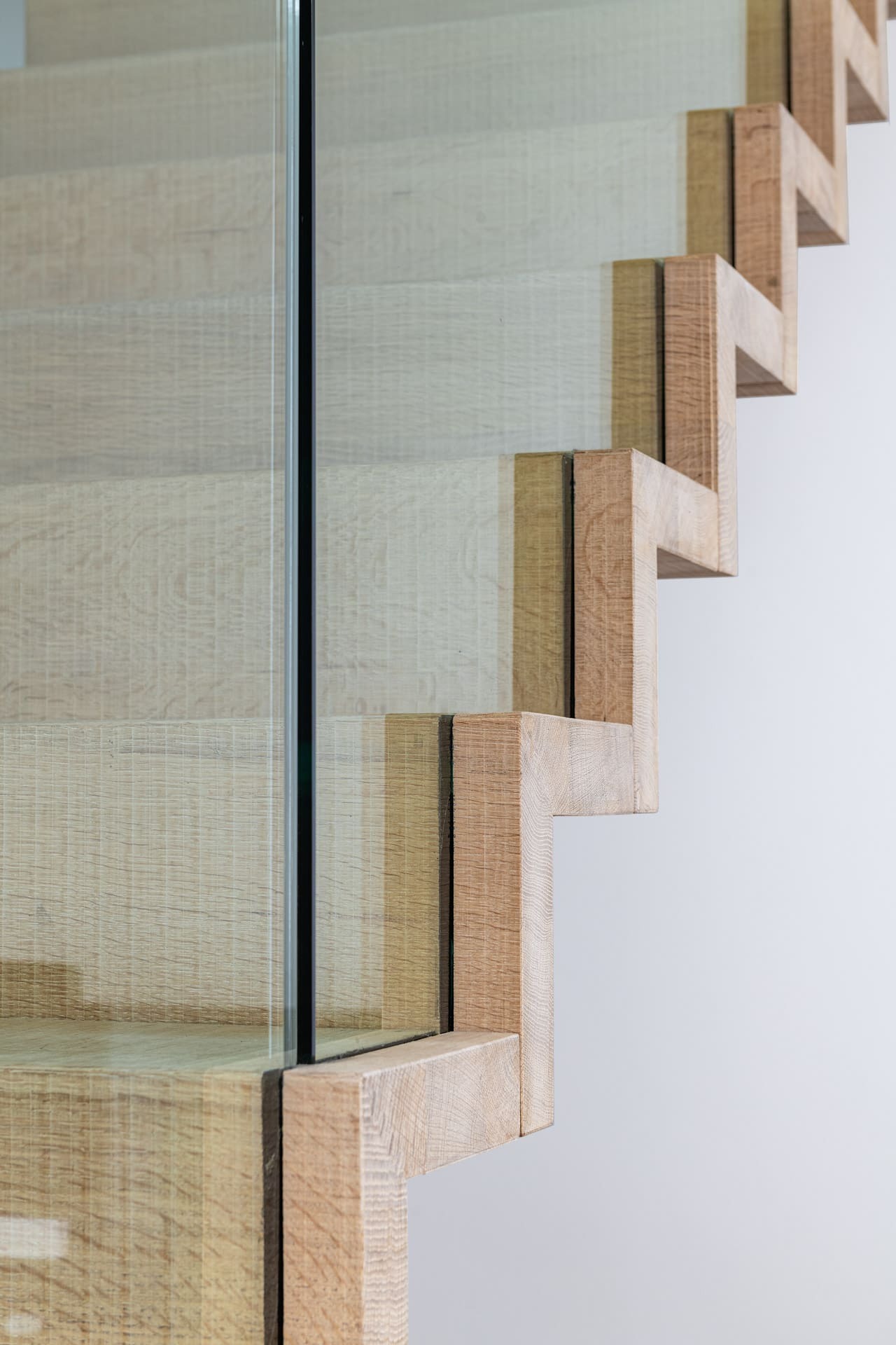 Close-up van een moderne trap met lichte houten zigzagtreden en een glazen balustrade, geplaatst in een strak afgewerkt interieur met witte muren.