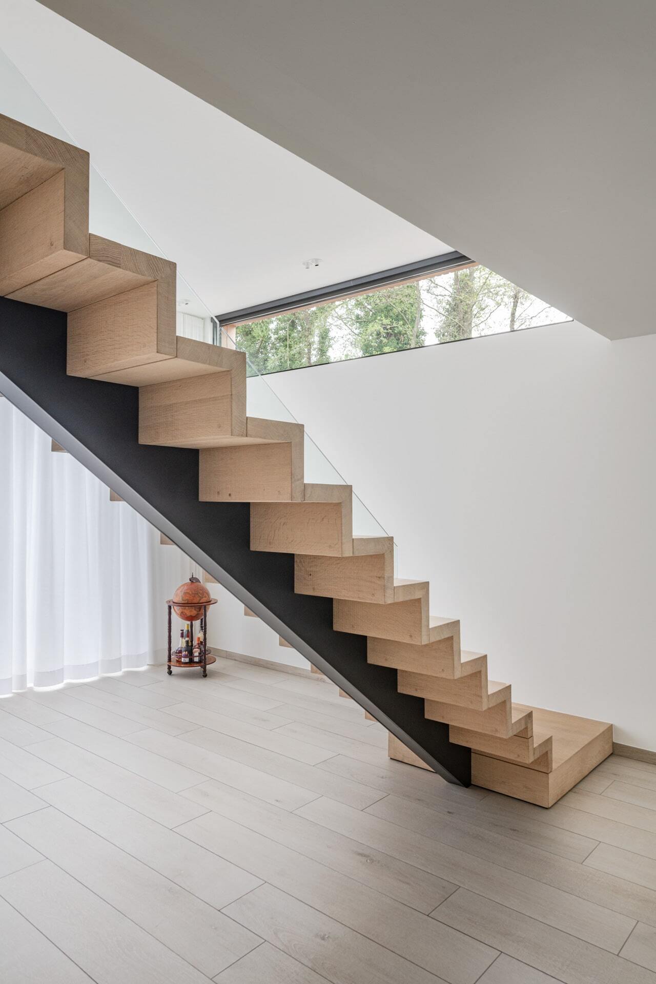 Moderne houten zigzagtrap met een zwarte stalen onderzijde in een minimalistische ruimte met lichte vloer, witte muren, gordijnen en een smal horizontaal raam.